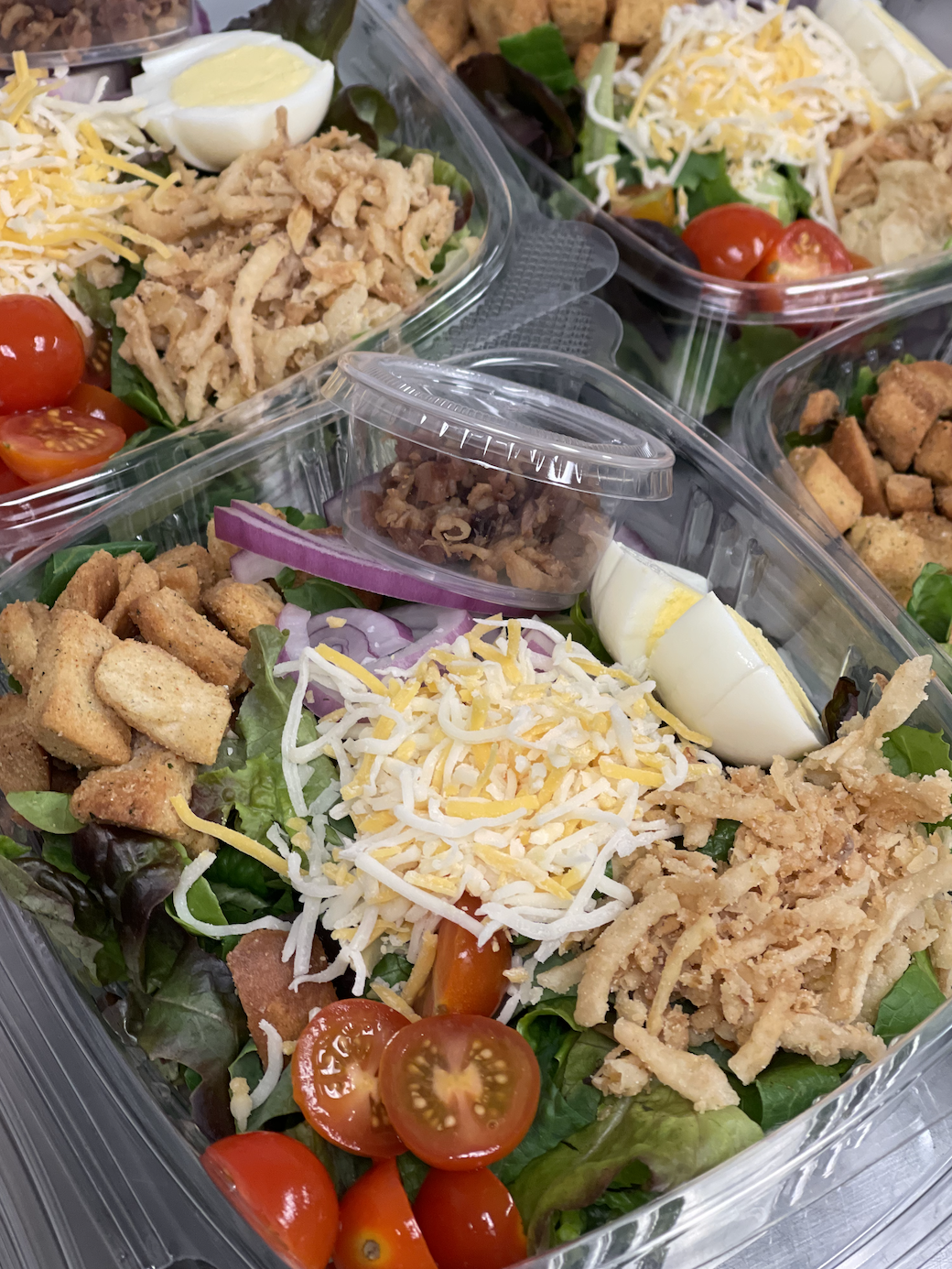 Container of salad with cherry tomatoes, shredded cheese, sliced red onions, chopped chicken, sliced boiled eggs, and chopped bacon.