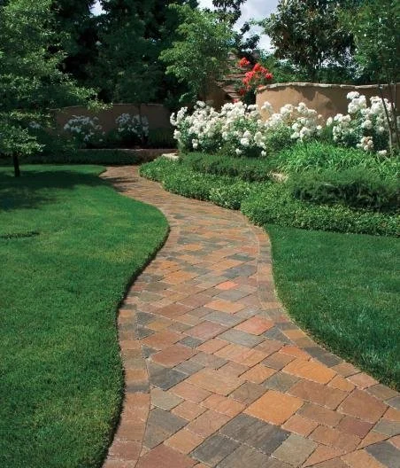 Curved brick pathway through a well-manicured garden with green grass, flowering bushes, and trees in the background.