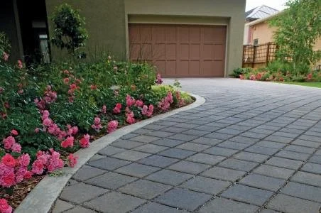 Paved driveway with brick border and flower beds with pink and red flowers, front of a house with a brown garage door.