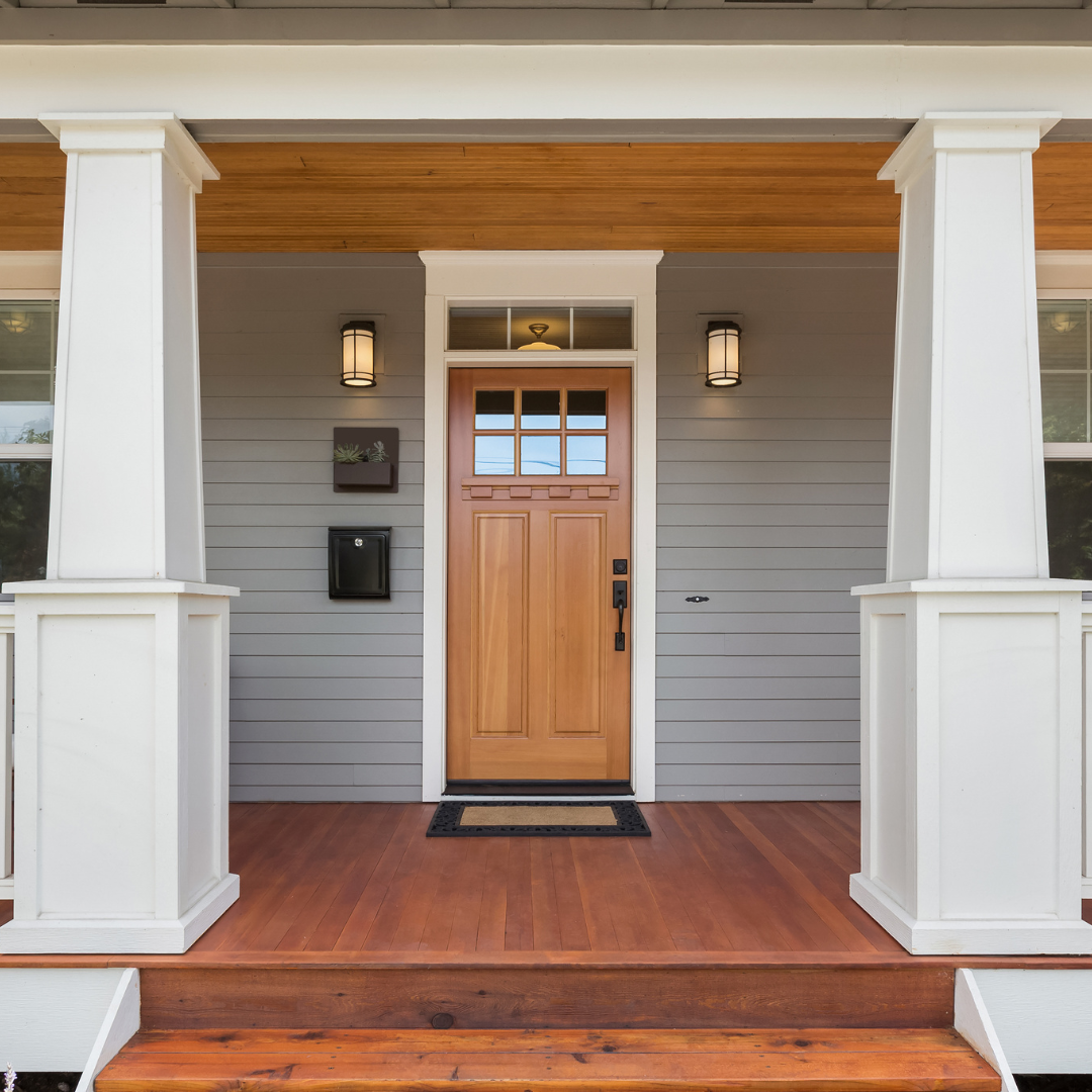 Front porch of a house with a wooden door, gray siding, two wall-mounted lights, a mailbox, and a small decorative plant