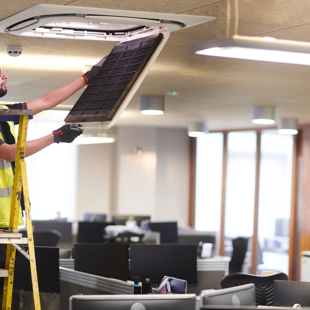 A technician installing a large ceiling vent or air conditioning unit in an office with multiple workstations and large windows.