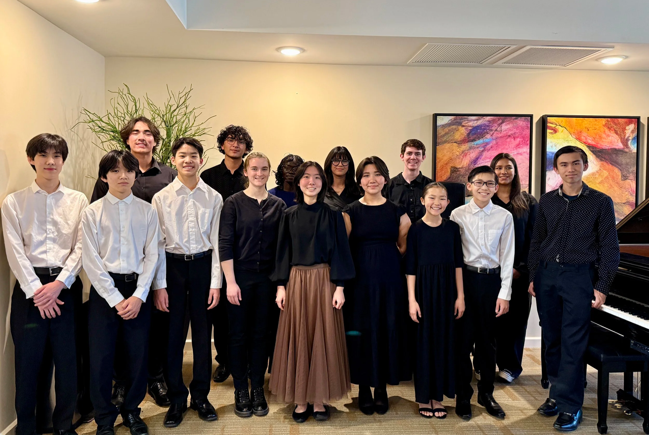 A group of 14 young people dressed in formal and semi-formal attire, standing indoors in front of colorful abstract art on the wall.