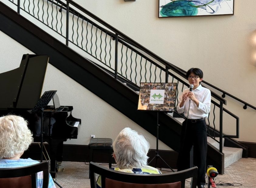 A young man standing next to a music stand, holding a microphone, speaking to an audience of older women seated in front of him, in a room with a staircase and a grand piano.