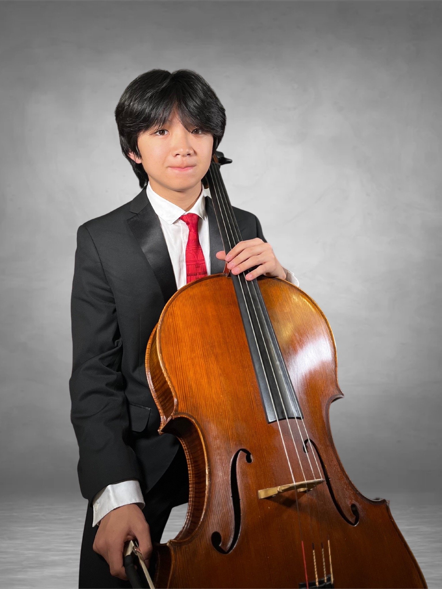Young boy in a black suit, white shirt, and red tie holding a large wooden cello against a plain gray background.