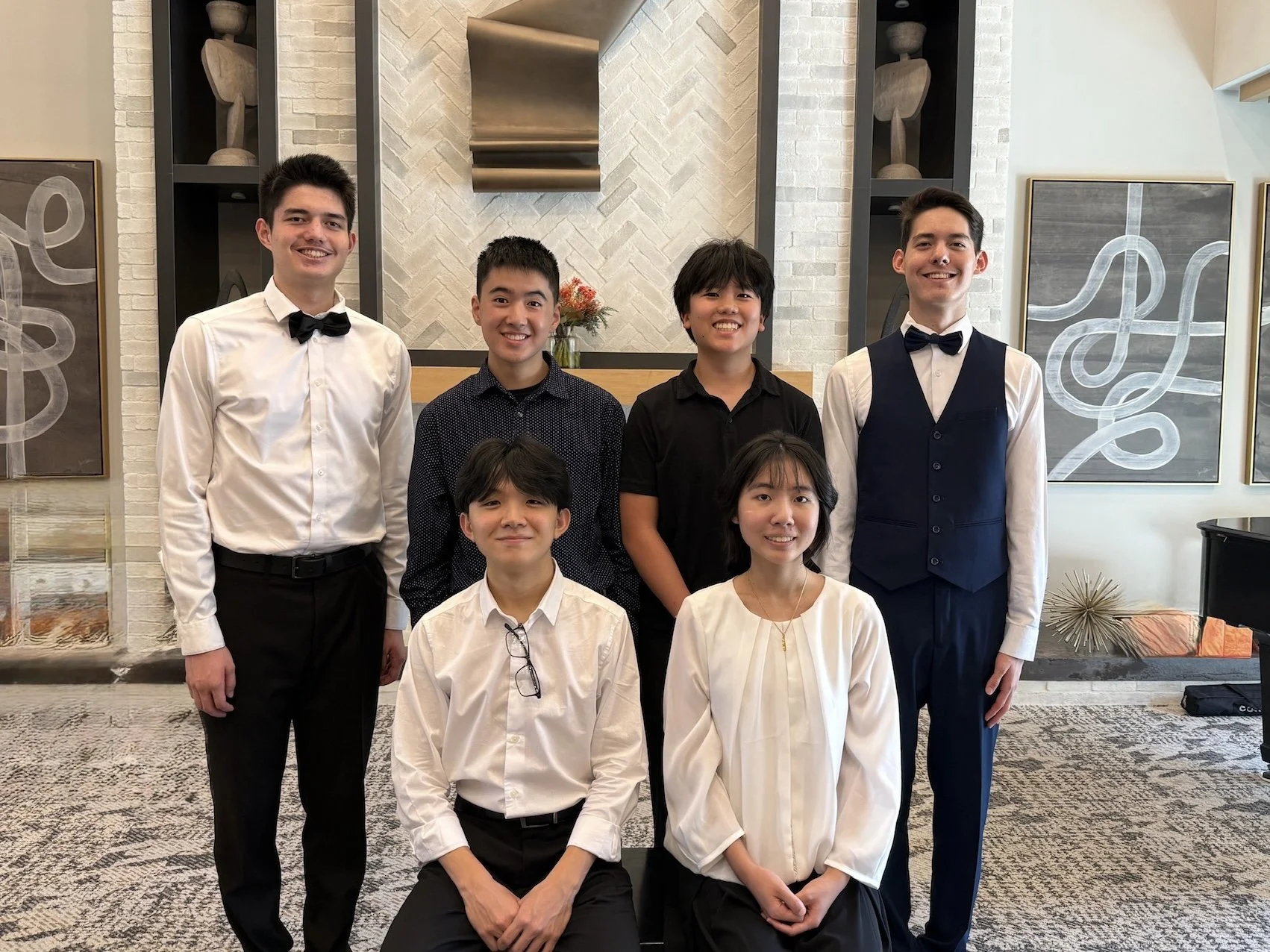 Group of six young adults dressed in formal attire indoors, smiling at the camera, with modern art and decorative objects on the wall behind them.