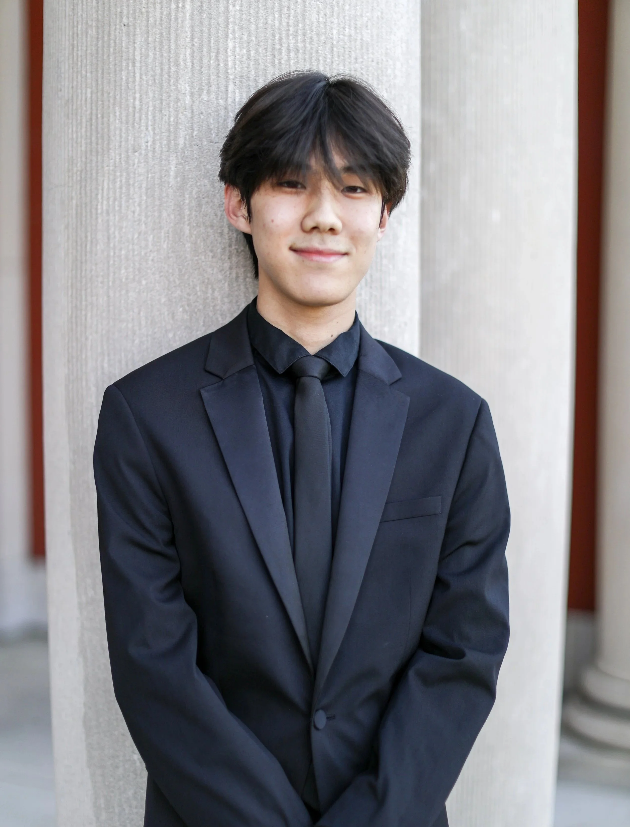 A young man in a black suit and black tie standing outdoors next to a light-colored column, smiling softly at the camera.
