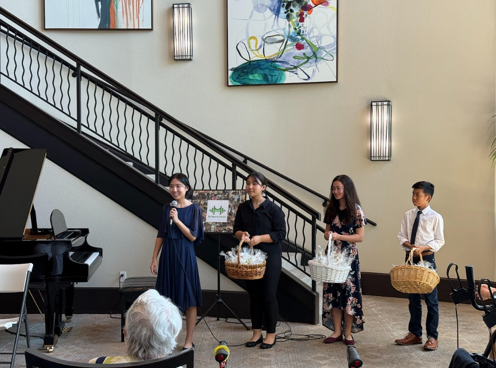 Four people standing in a line, with a woman holding a microphone, and three others holding baskets, in a room with a grand piano, a staircase, and colorful artwork on the wall.