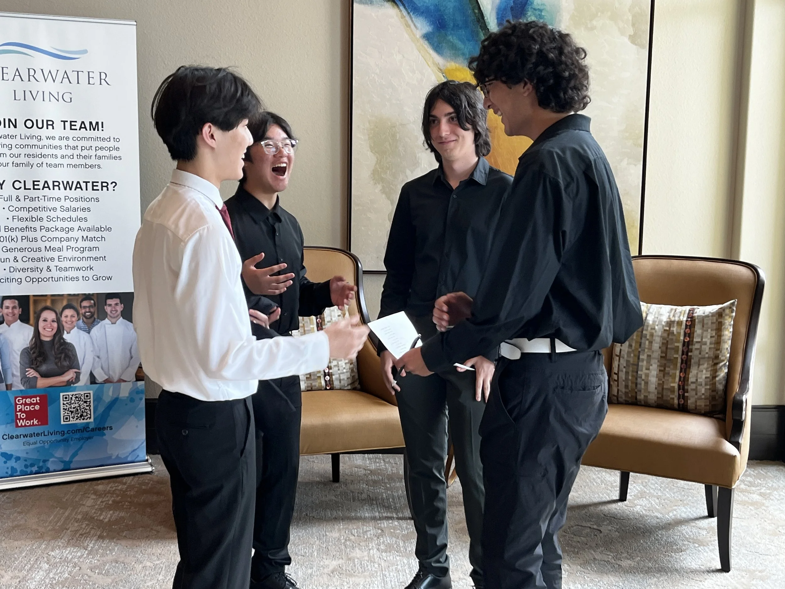 Four young men engaged in a lively conversation indoors near a promotional display for Clearwater Living, with a beige sofa and colorful abstract art on the wall behind them.