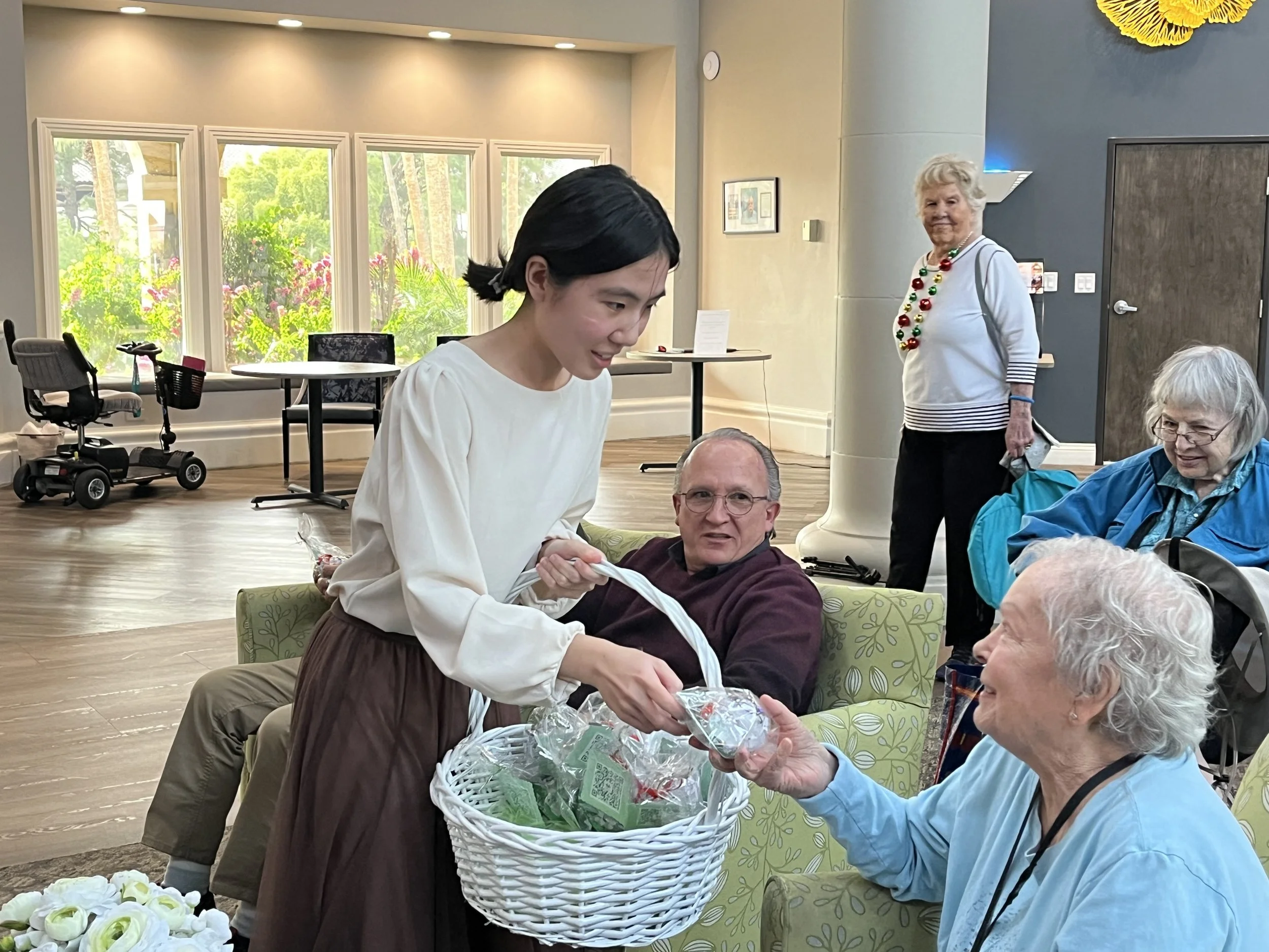 A young woman handing out wrapped items from a white basket to elderly women sitting in a common area of a care facility or community center, with other elderly women and a standing woman in holiday-themed jewelry in the background.