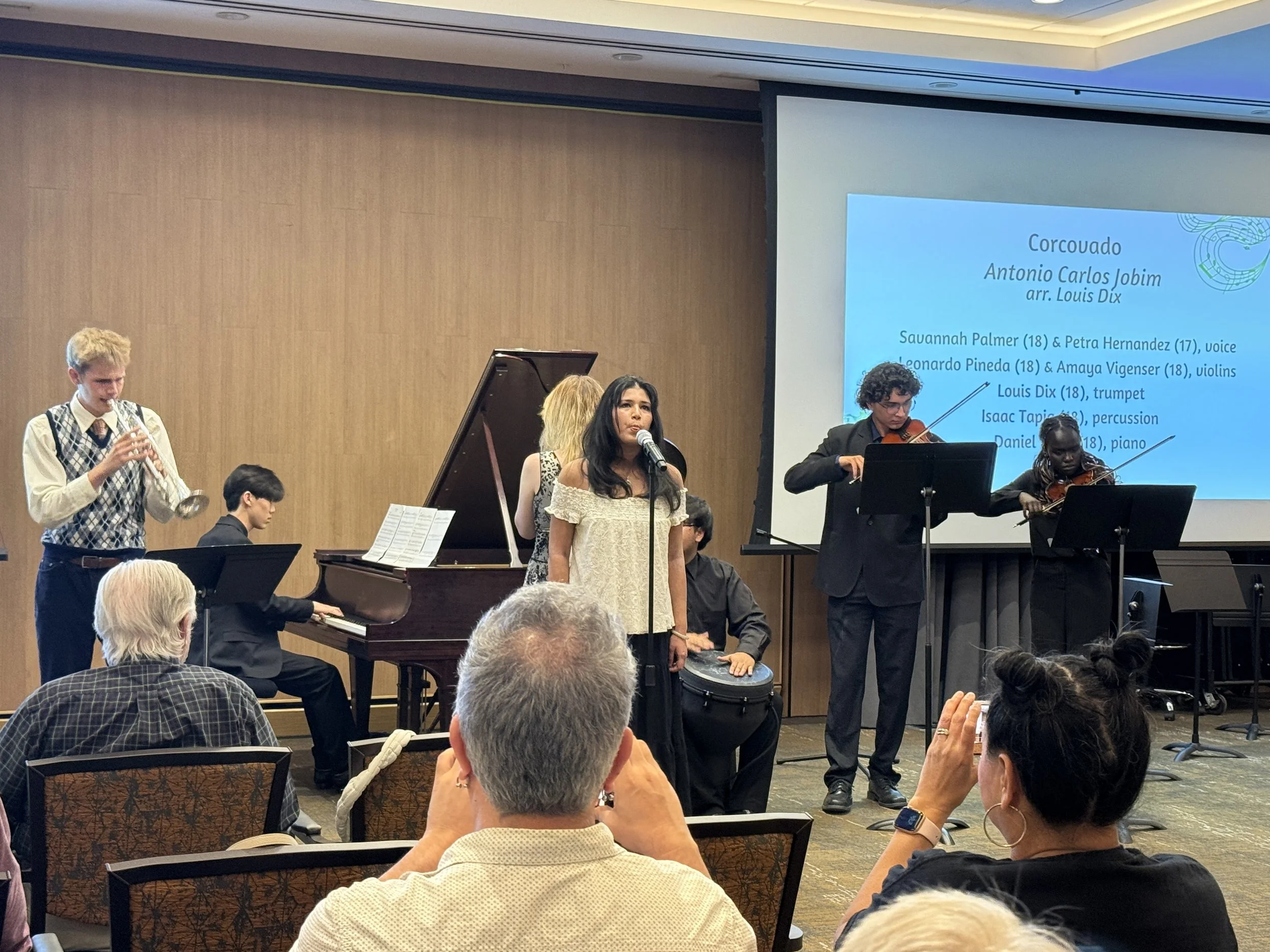 Group of young musicians performing on stage at a concert, including a violinist, a pianist, a flutist, and a singer, with an audience watching.