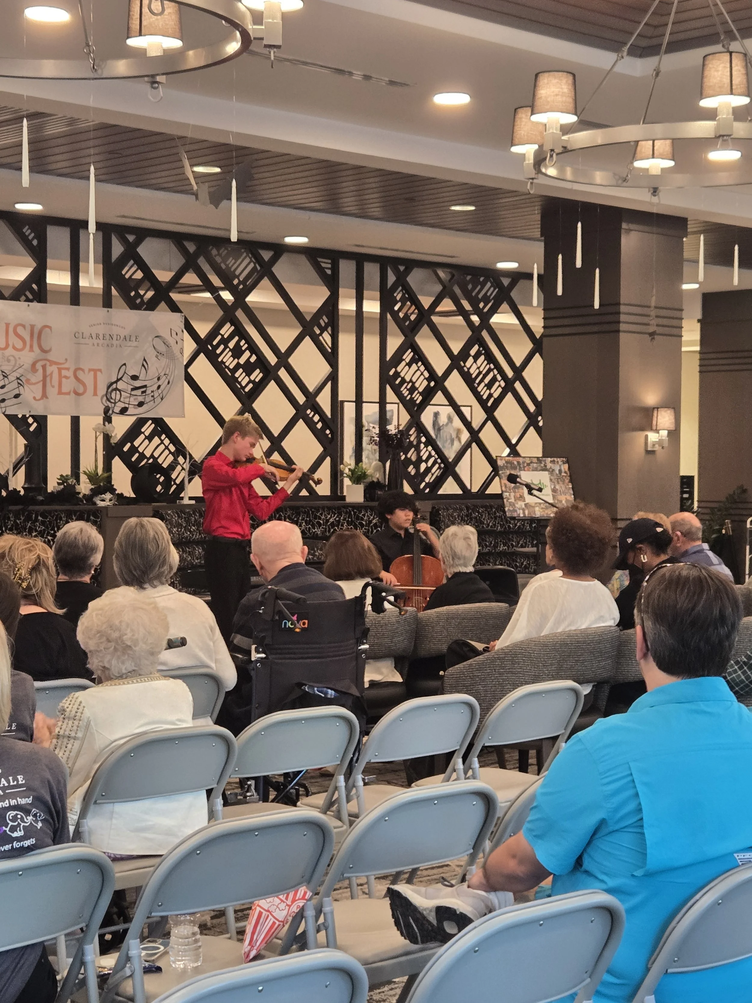 Young musician playing the violin and cellist performing in front of an audience at a music event in a decorated indoor venue.