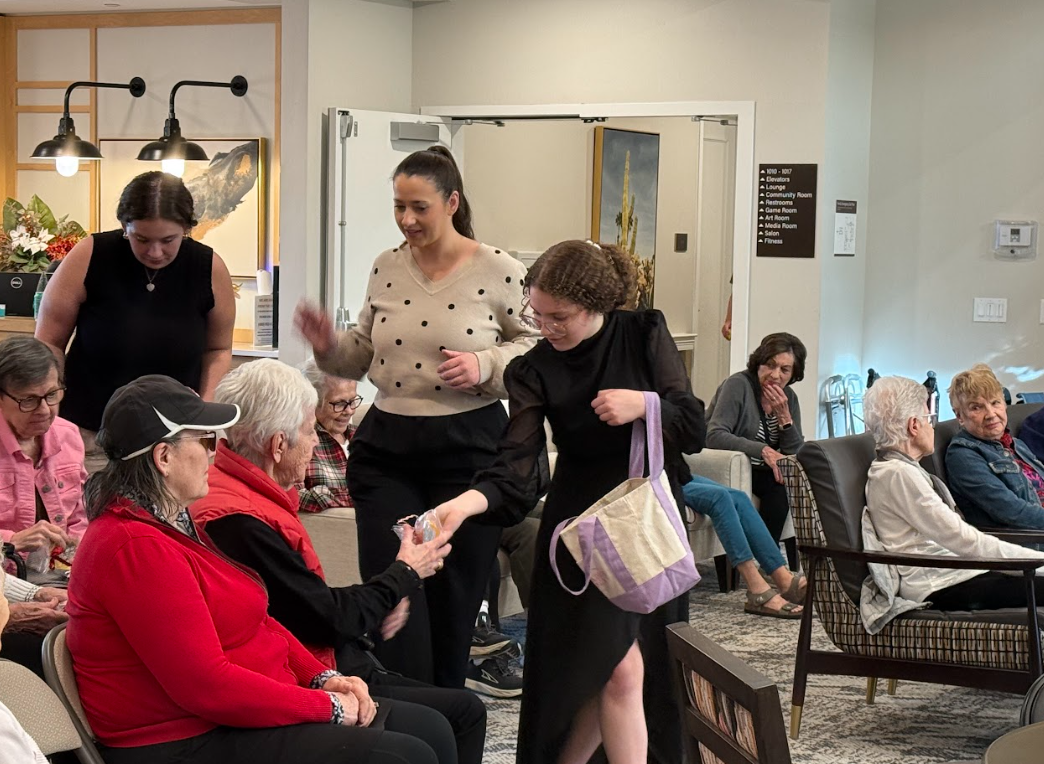 Group of women, mostly seniors, sitting in a lobby or communal room while three women approach and hand out small items, possibly gifts or snacks. The atmosphere appears relaxed and social in a community or care facility setting.