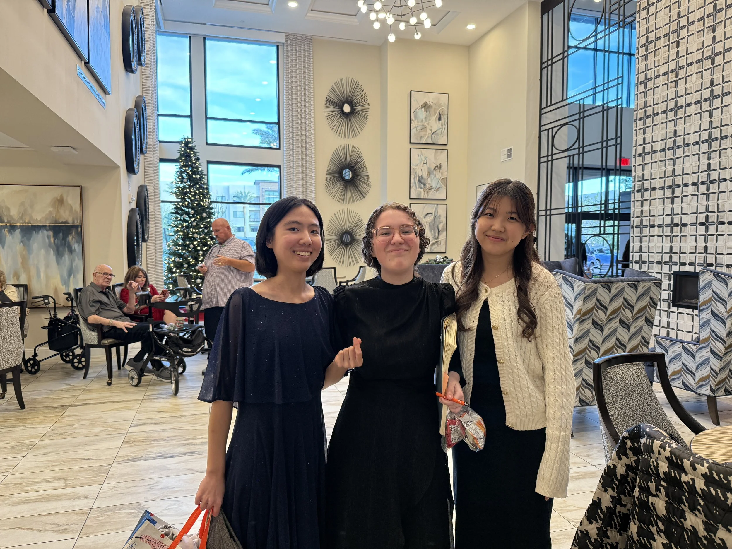 Three women smiling and posing together in a decorated indoor space with a Christmas tree in the background. Others are seated and standing nearby, with large windows and artistic wall decor.
