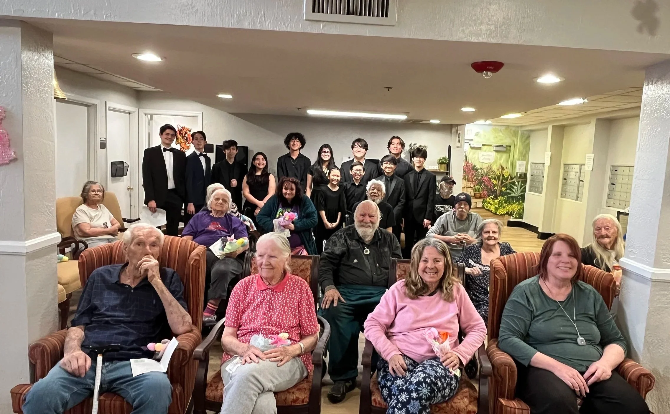 Group of elderly residents and staff at a care home celebrating Easter, with some holding colorful Easter eggs and flowers, and younger staff dressed in black formal attire standing in the back.
