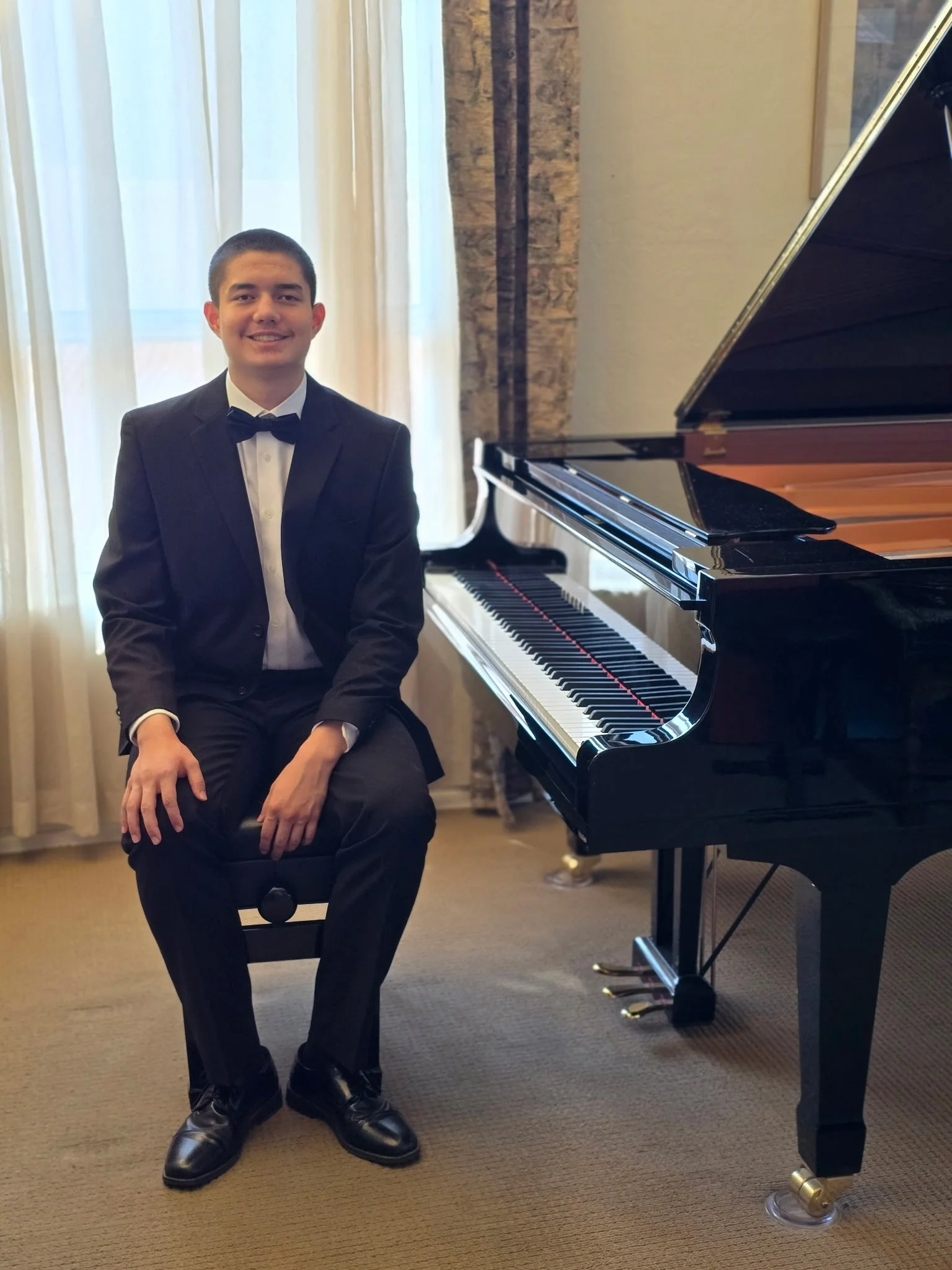 A young man in a tuxedo sitting on a stool next to a grand piano in a well-lit room with curtains and a brick wall.