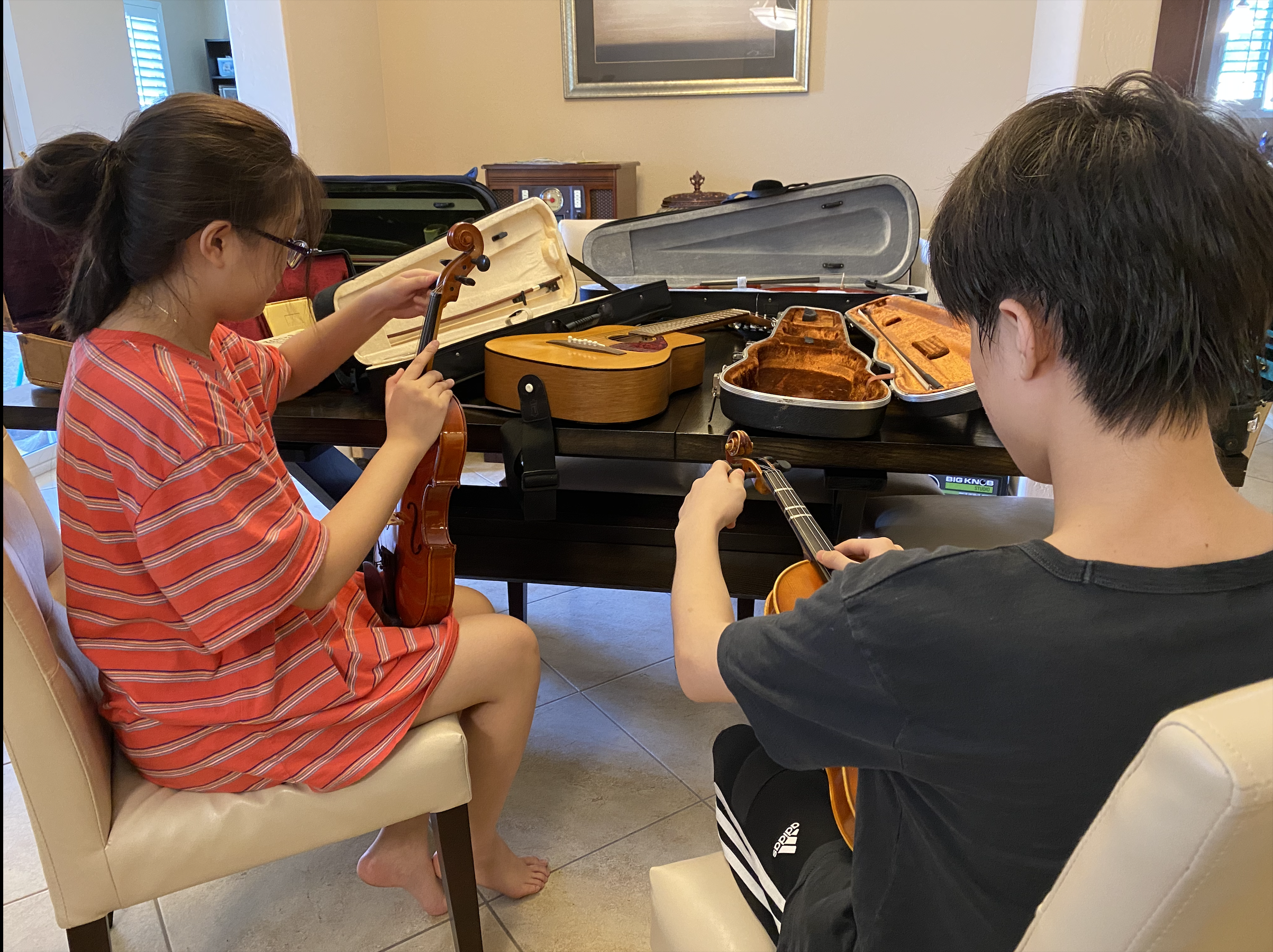 A young girl and boy sitting at a table, each holding a violin, surrounded by open violin cases in a room.