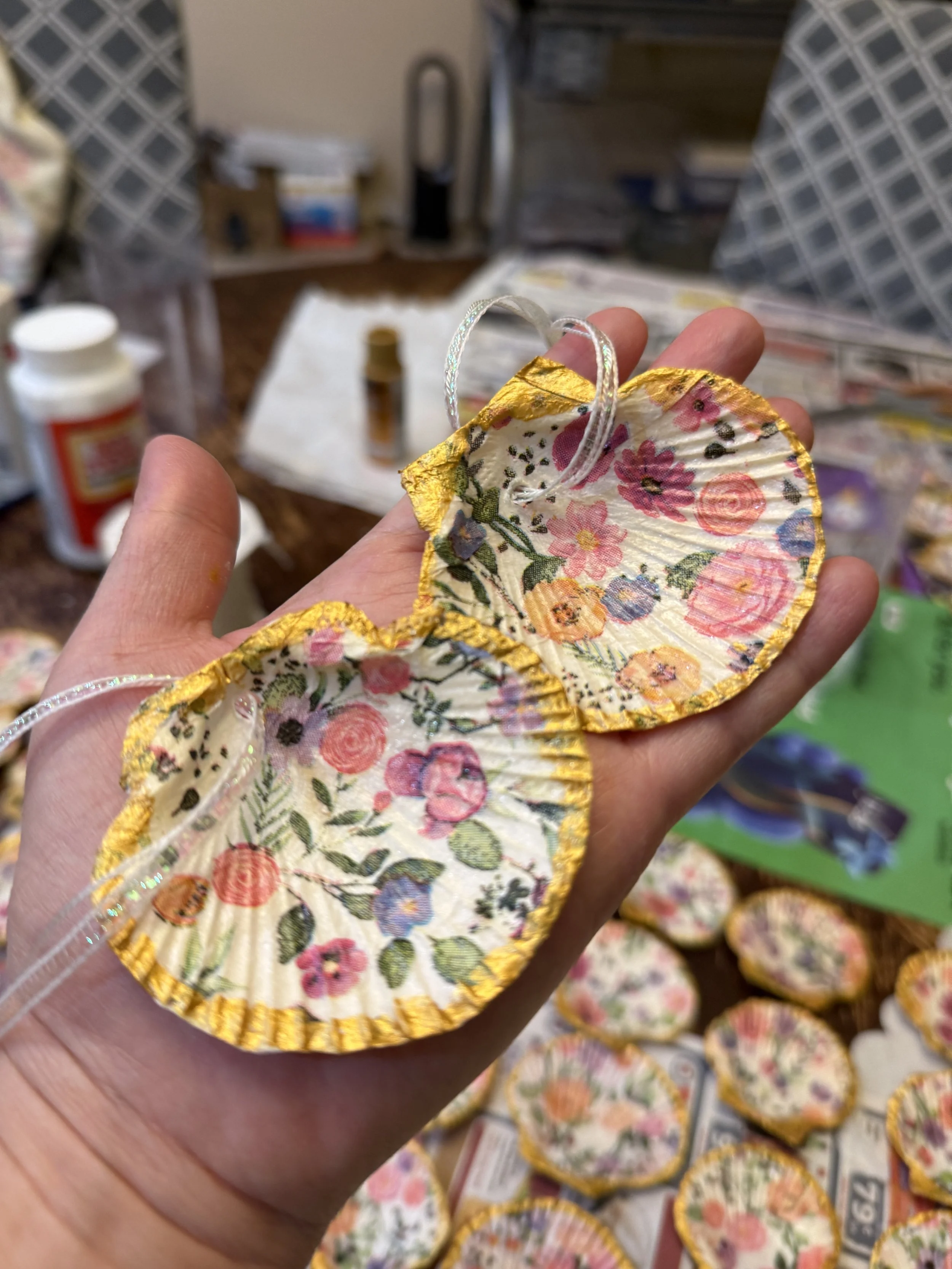 Hand holding two shell ornaments with floral patterns and yellow borders, surrounded by more similar ornaments on a table in a craft room.