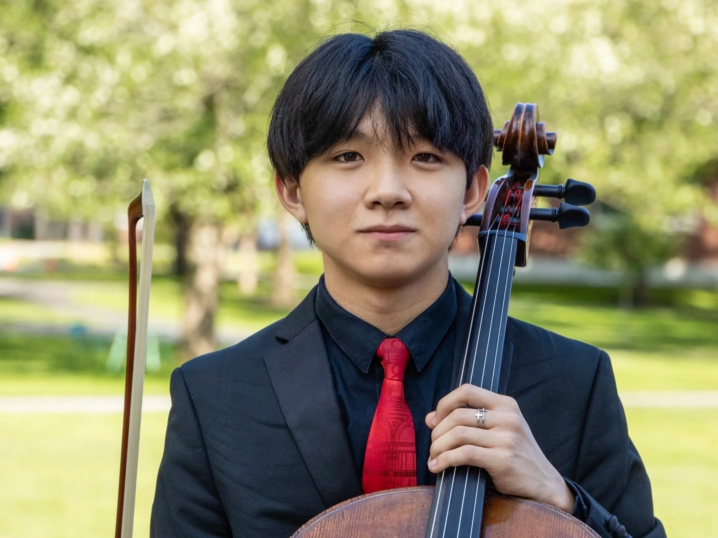 A young person with black hair and light skin holding a violin and bow, dressed in a black suit and red tie, standing outdoors with a background of green trees and grass.