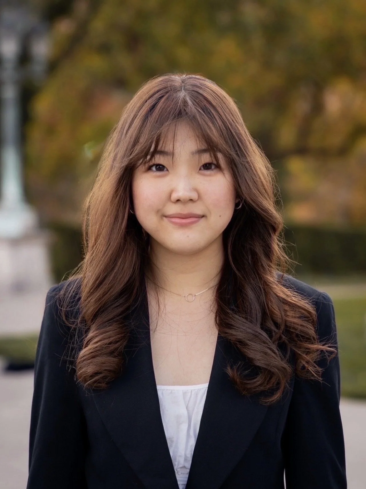Portrait of a young woman with long brown hair, wearing a black blazer and a white top, standing outdoors with blurred autumn trees in the background.
