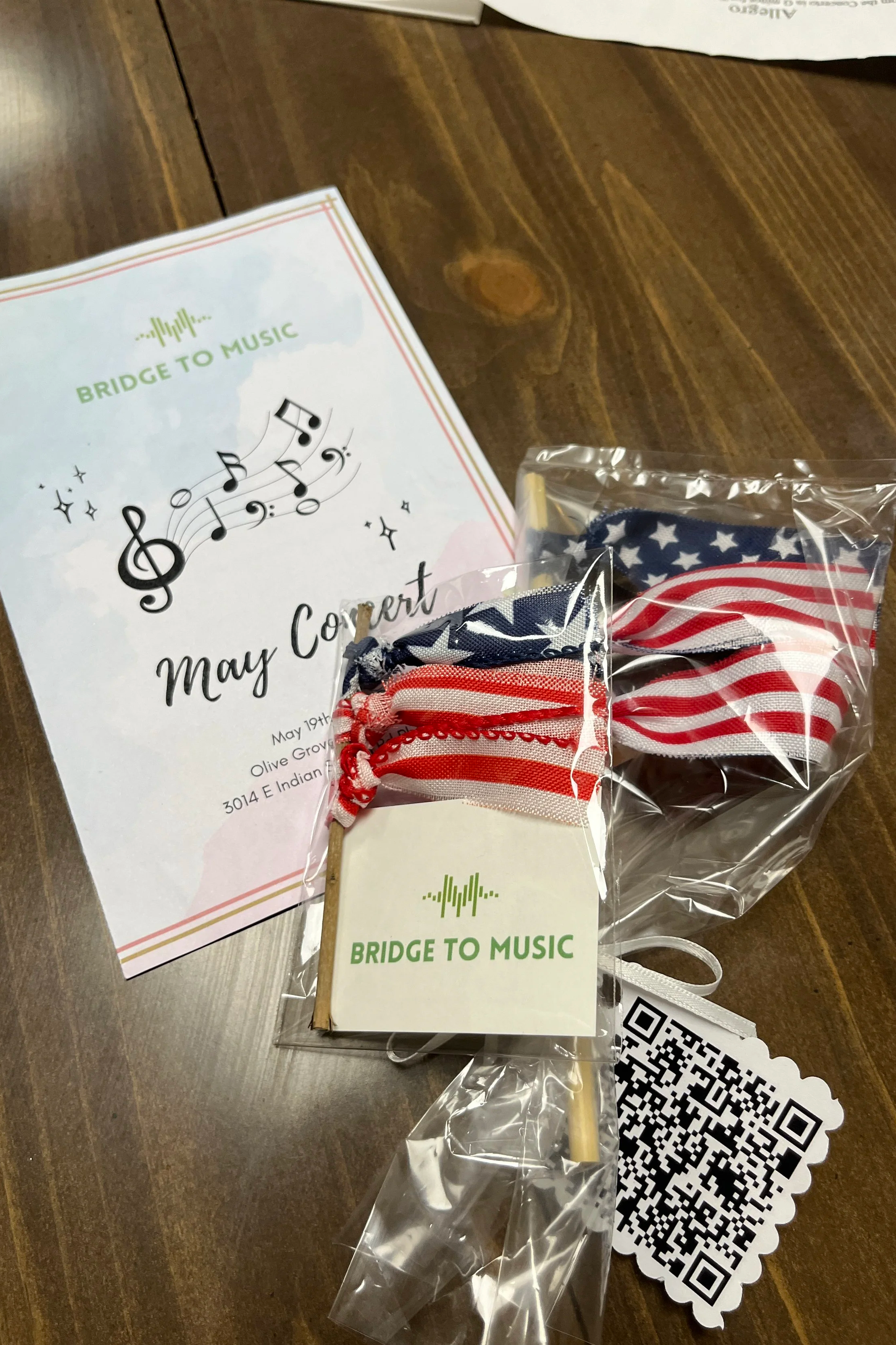 A May concert flyer, American flag-themed ribbons, a QR code, and a 'Bridge to Music' sticker on a wooden table.