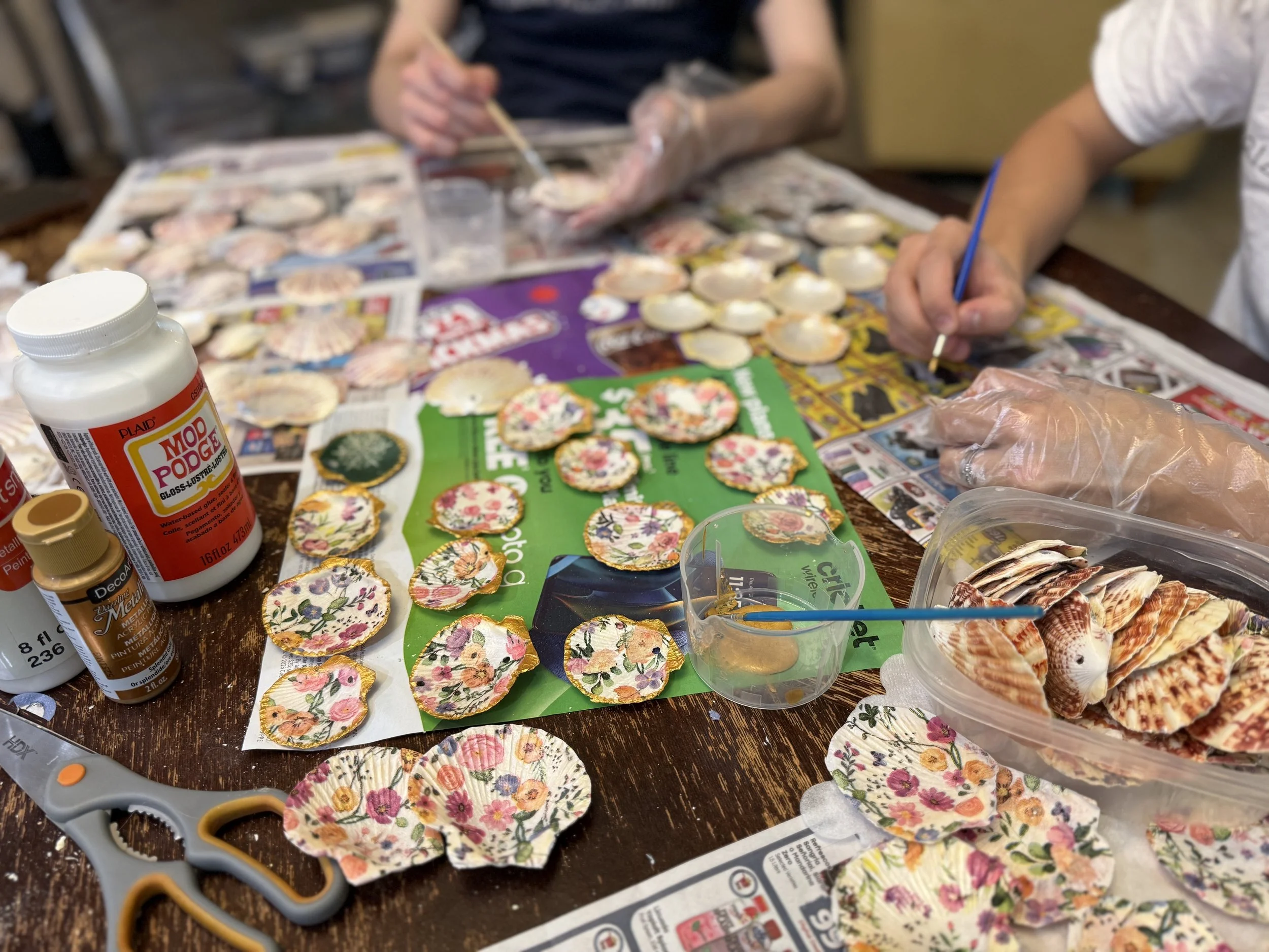 People decorating seashells with floral patterns, using paints and brushes on a table covered with newspapers and arts and crafts supplies.