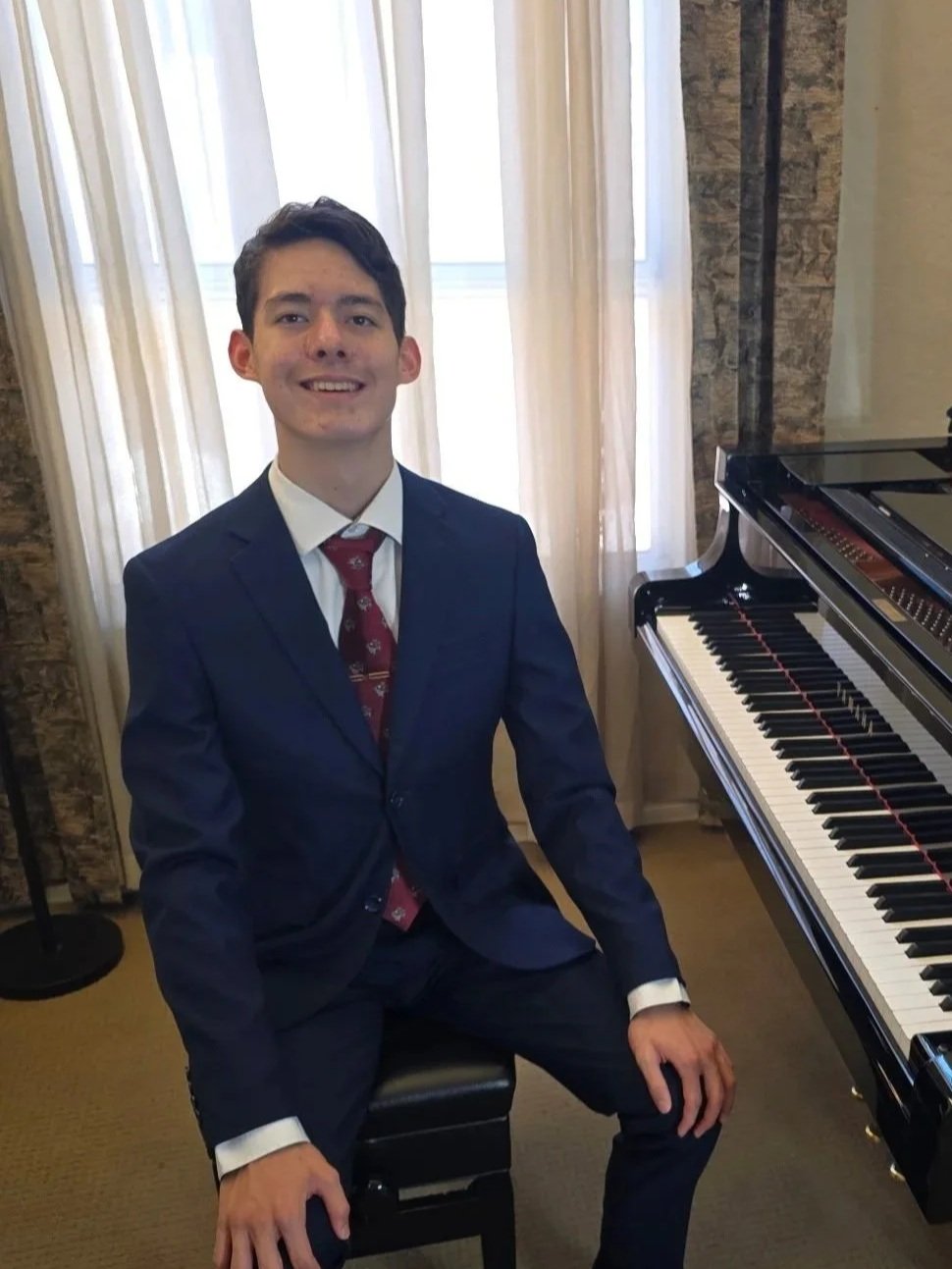 A young man in a navy suit and red tie sitting on a black piano bench next to a grand piano, smiling at the camera in a room with beige curtains and stone walls.