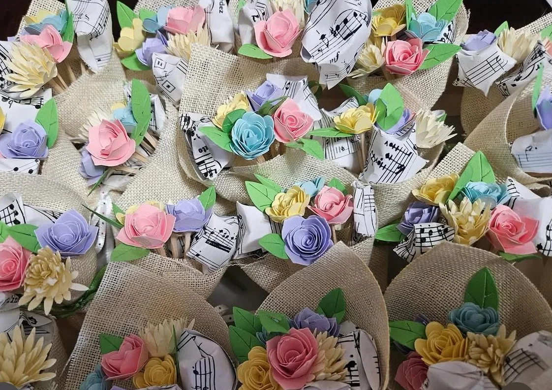 Multiple handmade paper flower corsages with colored flowers and sheet music paper accents, arranged on a surface.