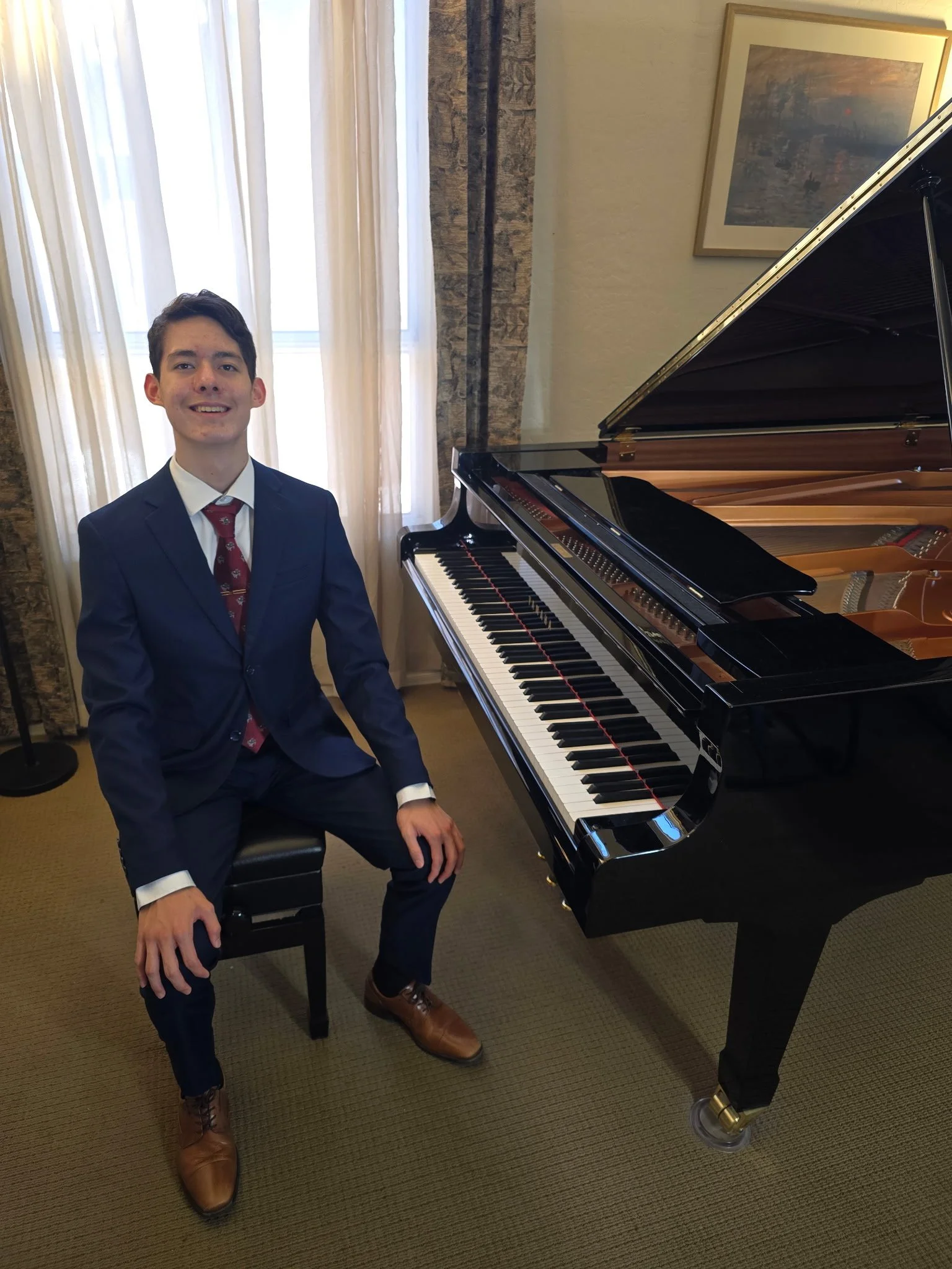 Young man in a suit sitting beside a grand piano in a well-lit room with curtains and artwork on the wall.
