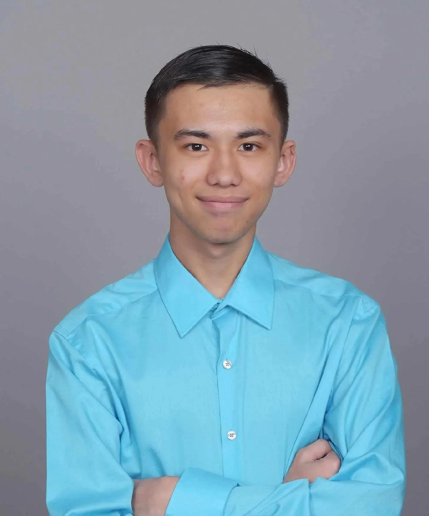 Young man with short dark hair, wearing a bright blue button-up shirt, standing against a plain gray background, with his arms crossed and smiling slightly.