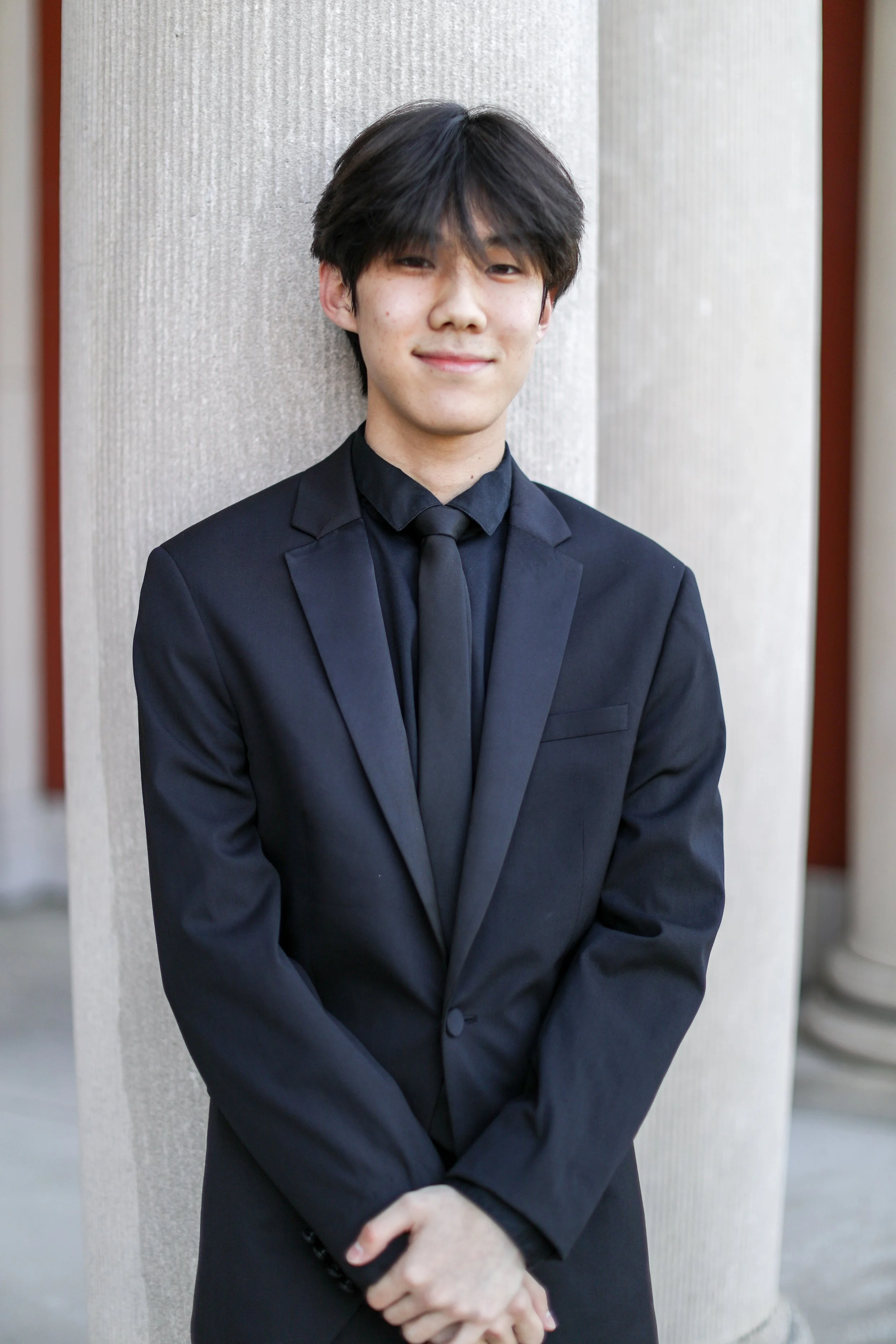 Young man in a black suit and tie standing outside, smiling with arms crossed, leaning against a concrete column.