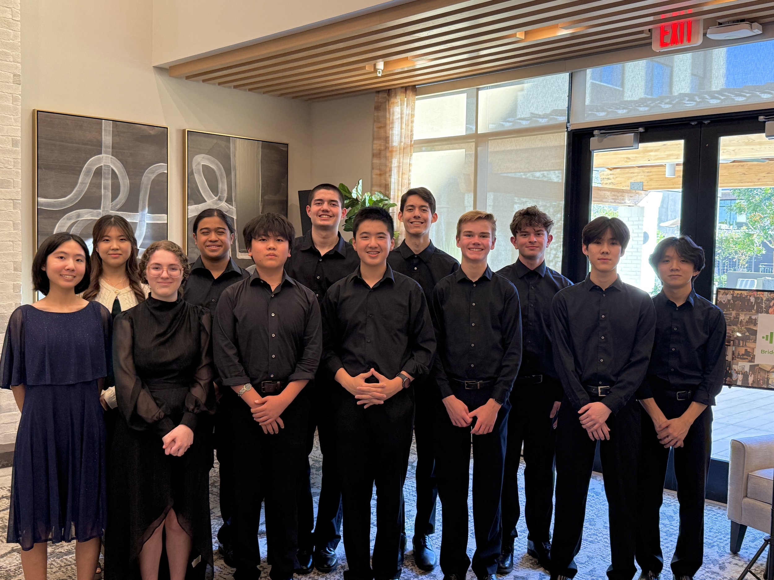 Group portrait of eleven young people, some smiling and some serious, dressed in black and dark clothes, standing inside near glass doors with artwork on the wall behind them.