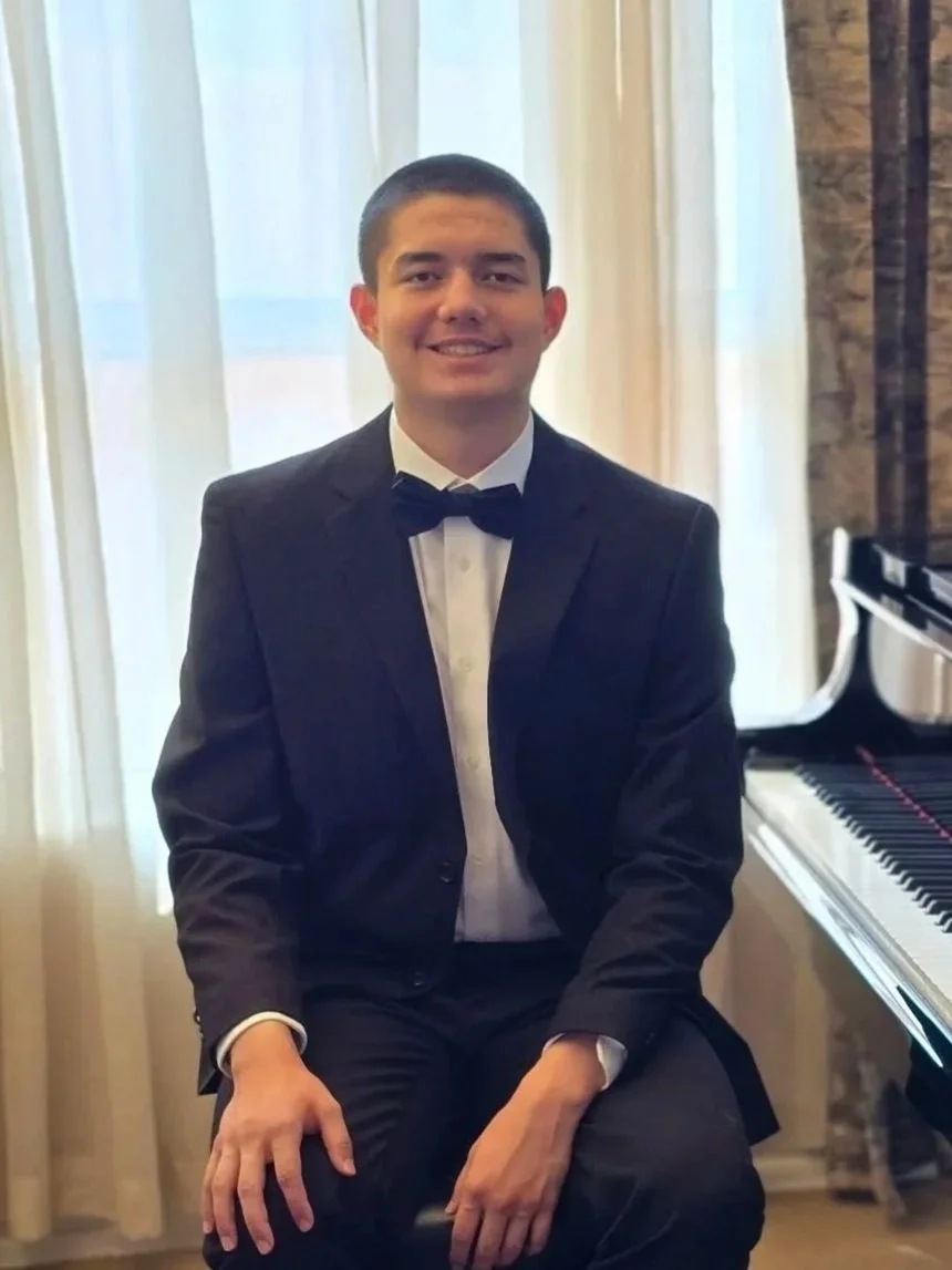 A young man in a tuxedo sitting next to a grand piano in a room with light-colored curtains.