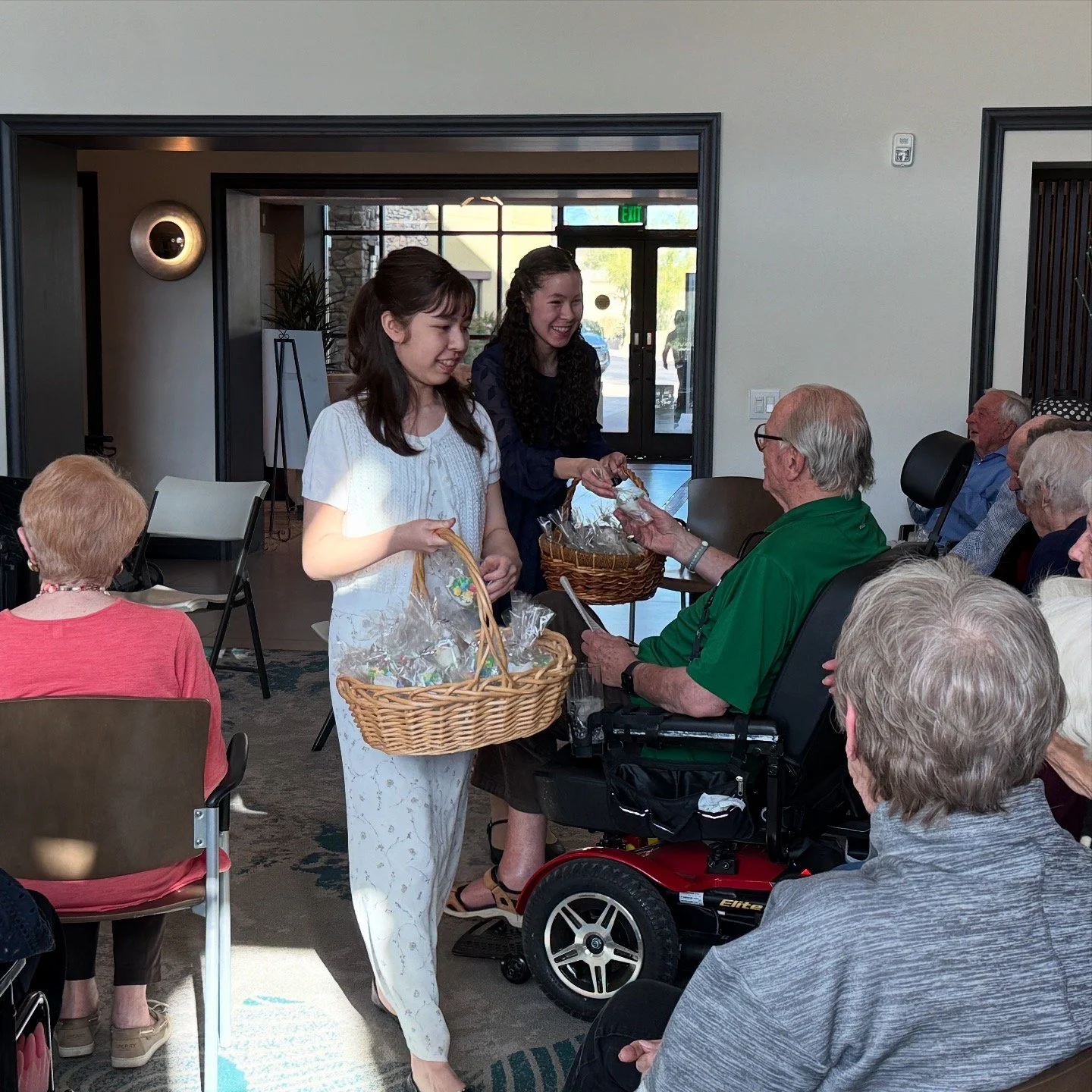 Two young women handing out small gift baskets to elderly residents in a common room, surrounded by seated elderly people, some in wheelchairs, in a social gathering at a care facility.