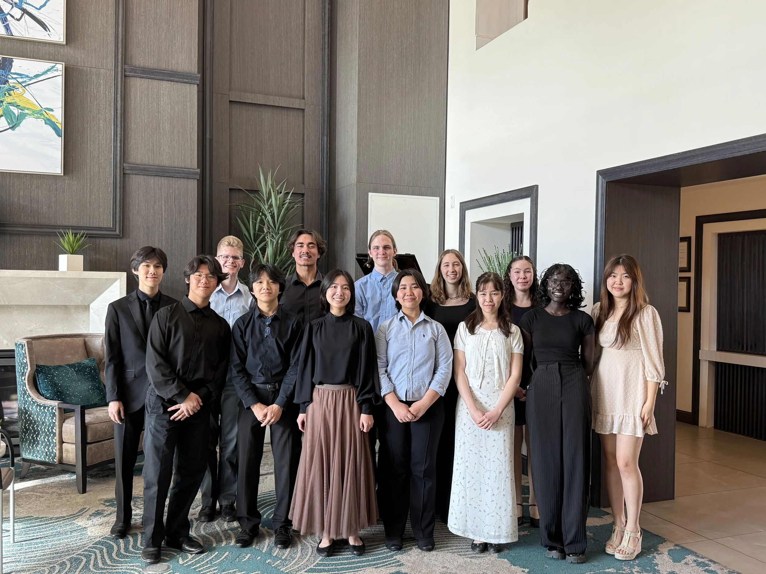 Group of fifteen young adults posing indoors in formal attire, standing in front of a dark wood wall with artwork and plants, with a white wall and door frame visible.