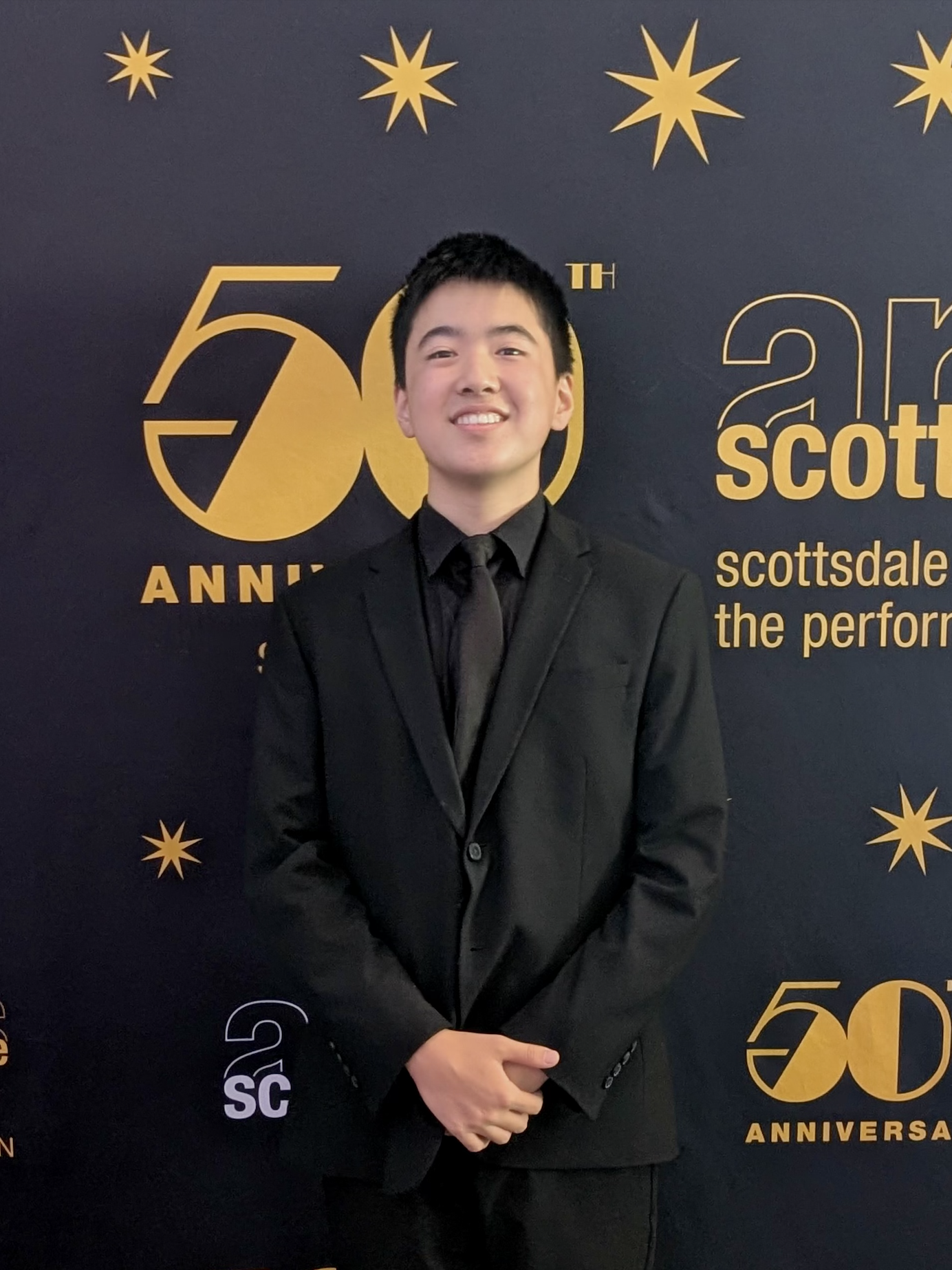 A young man in a black suit smiling, standing in front of a black backdrop with gold stars and text that says '50th Anniversary' and 'scottsdale the perform'.