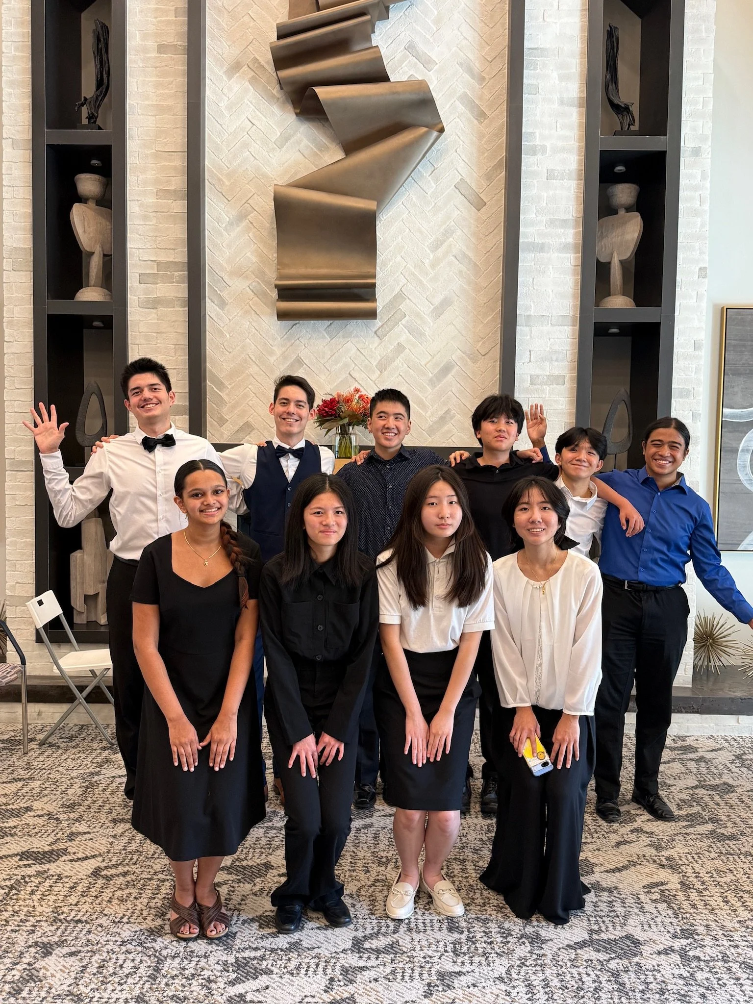 A group of young adults dressed in formal and semi-formal attire, standing in front of a modern, stylish background with white brick wall and decorative sculptures. They are smiling, some with arms around each other, appearing to be at a celebratory or social gathering.