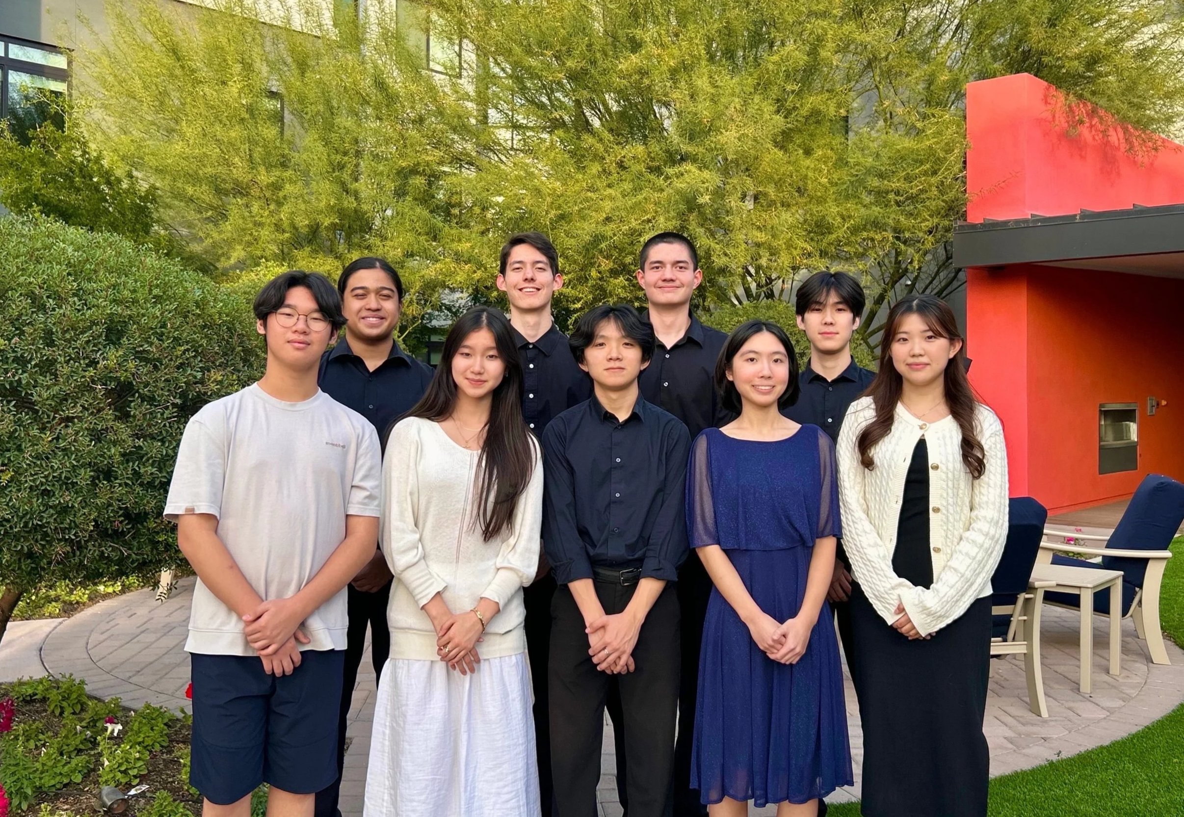 Group of nine diverse young adults standing outdoors on a paved pathway with green trees and bushes in the background. Some are smiling, dressed in casual and semi-formal attire, with a modern building and orange structure visible on the right side.