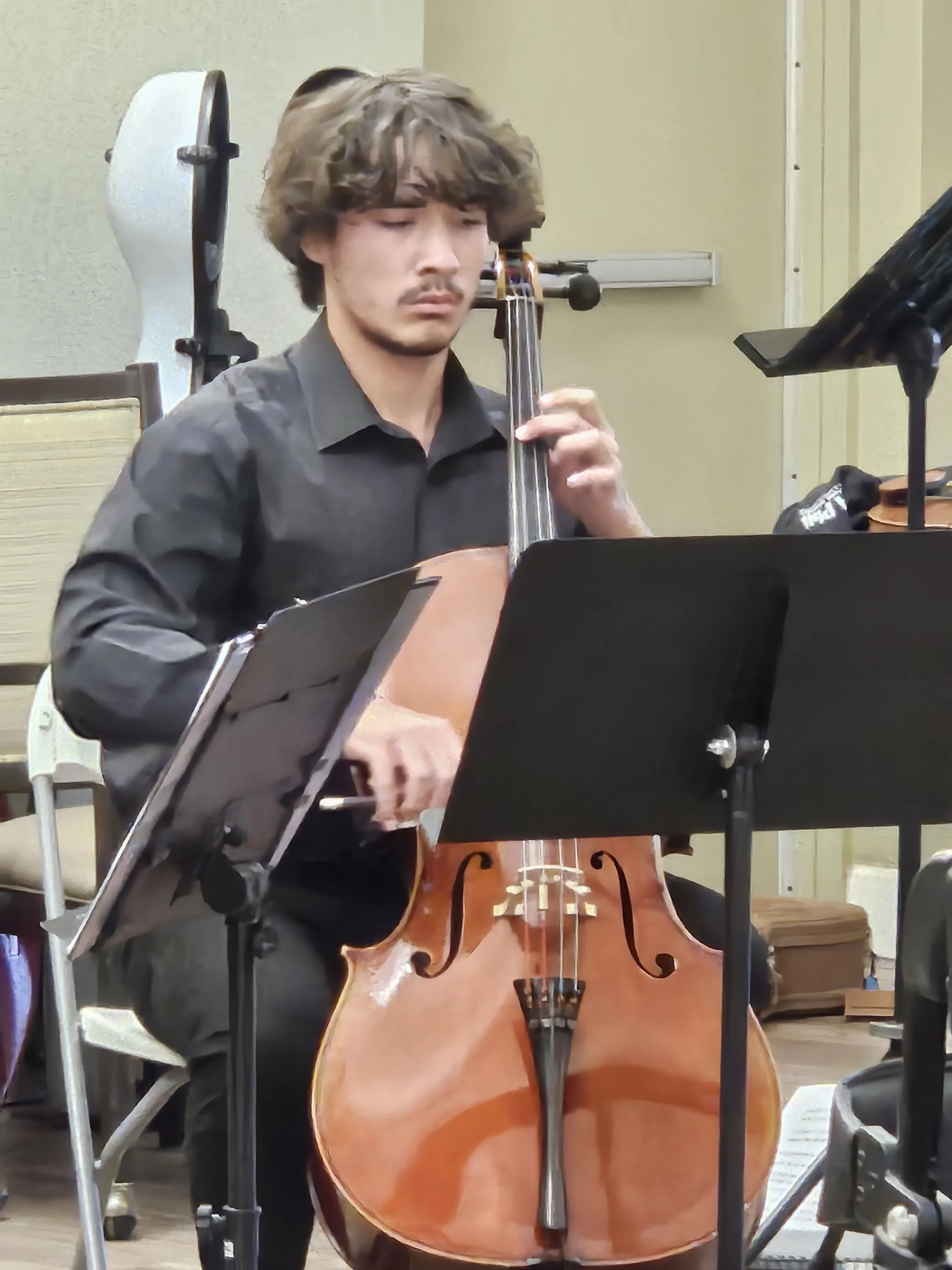 A young man with wavy brown hair playing a cello, wearing a black shirt, in a music rehearsal room with music stands and other musical equipment.