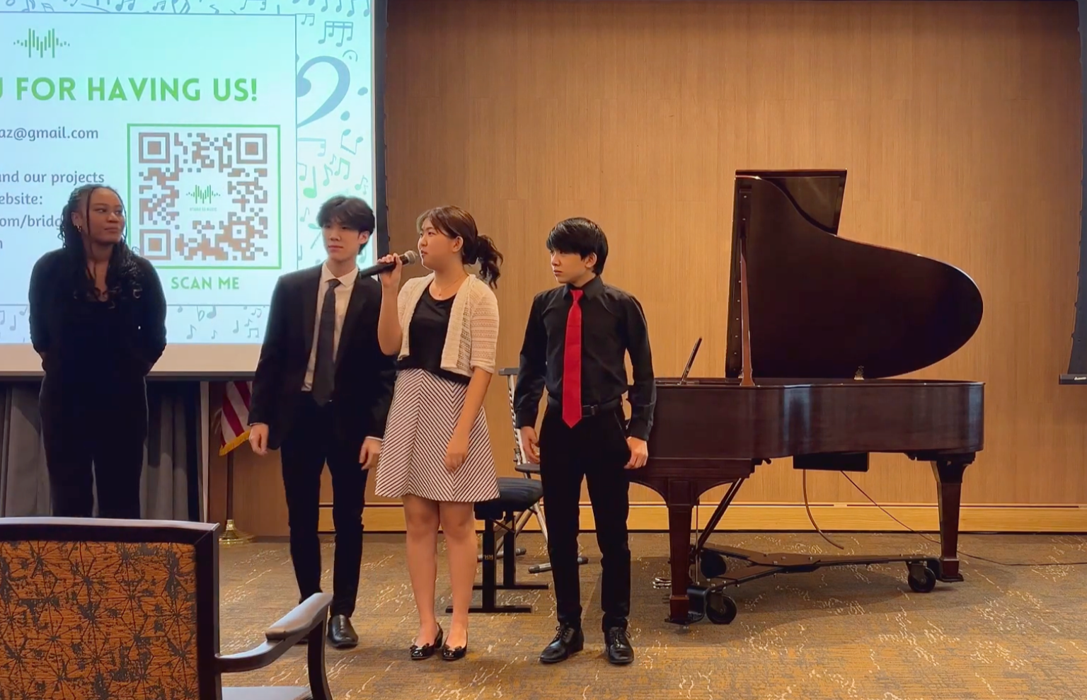 Four young people standing on a stage with a grand piano, two men in suits and a woman holding a microphone, while a woman is dressed in black. Behind them, a large screen displays a QR code and some text.