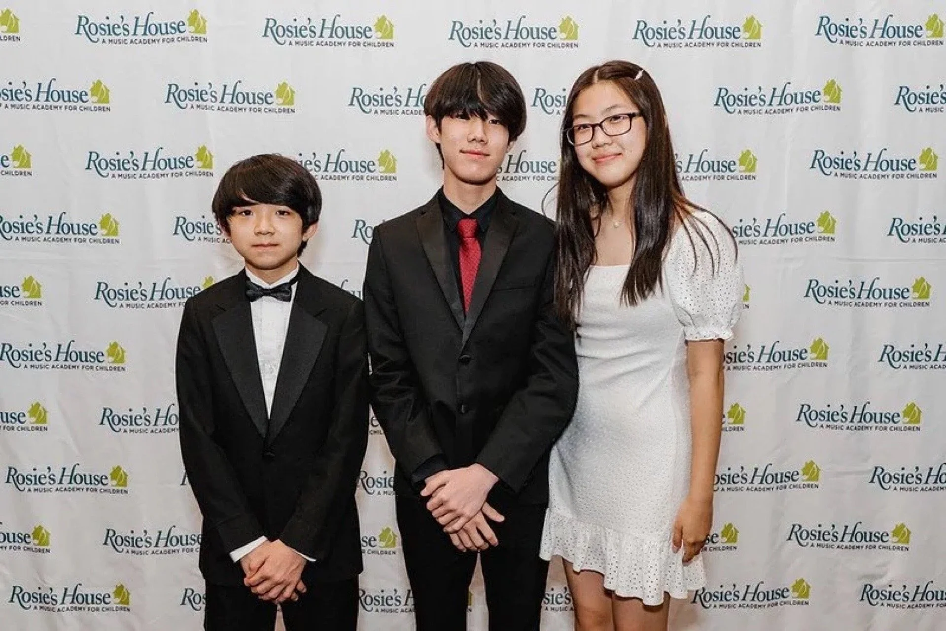 Three young people dressed in formal attire standing in front of a backdrop with 'Rosie's House' written repeatedly.