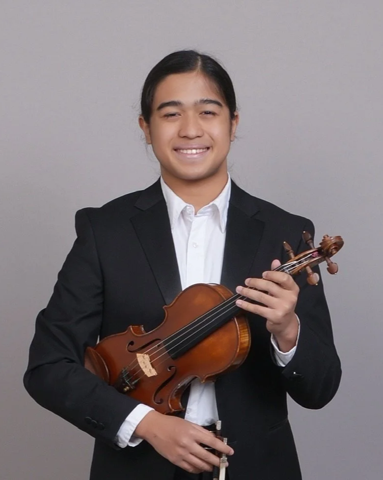 A young man in a black suit holding a violin and bow, smiling at the camera, standing against a plain gray background.