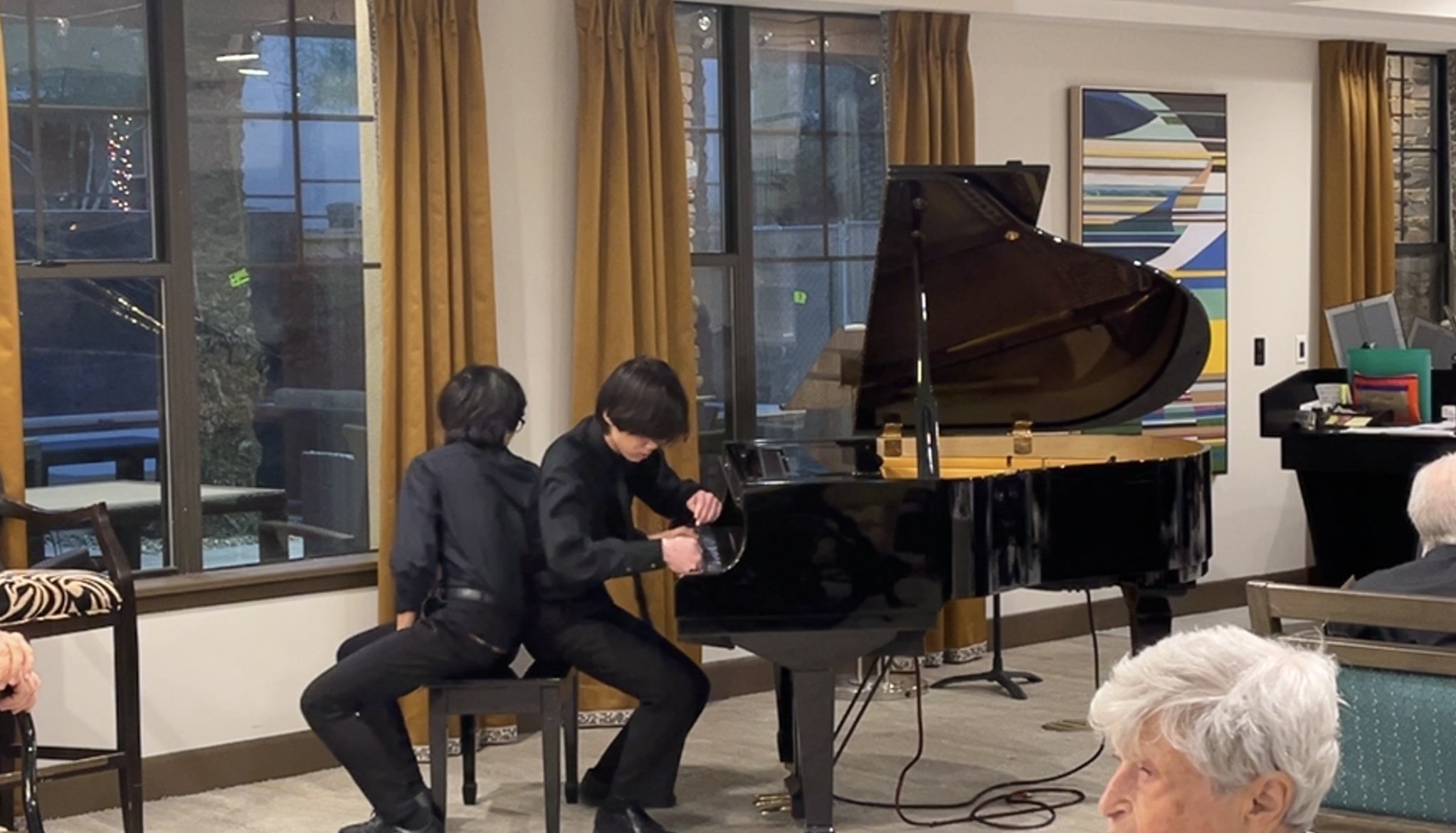 A young man with black hair and glasses playing a piano in a room with large windows and yellow curtains, with older people sitting and watching him.
