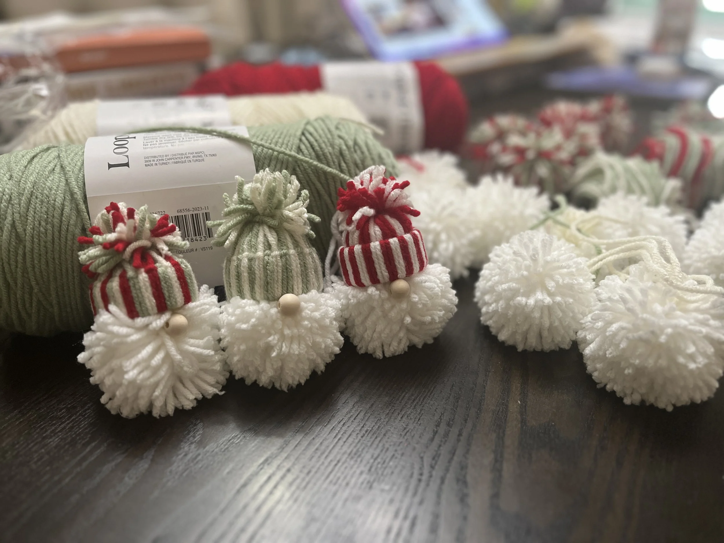 A collection of Christmas-themed pom pom garlands and yarn skeins on a wooden surface. The garlands have small knit hats with pom poms, featuring red, white, and green colors.