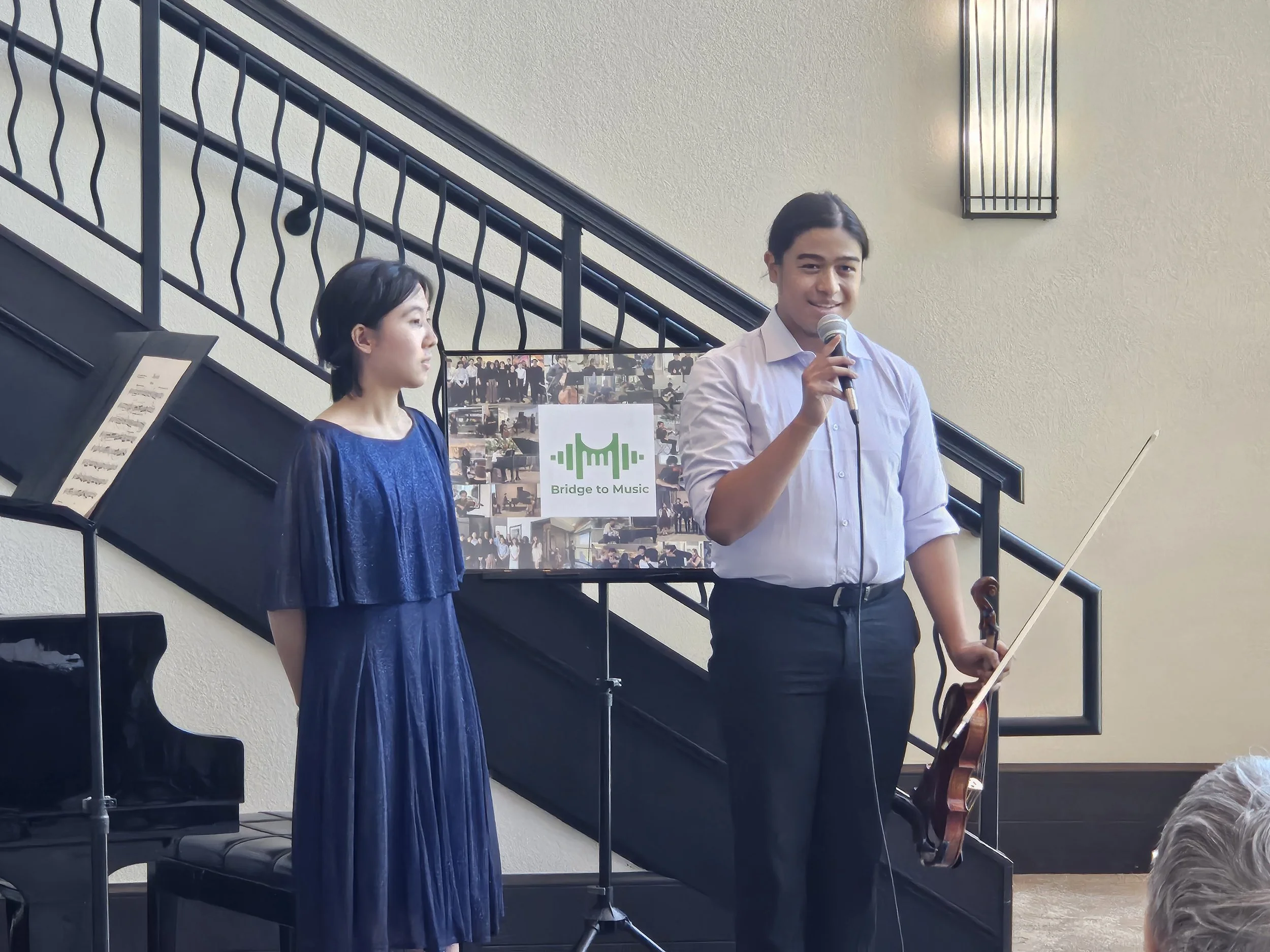 A young man holding a microphone and a violin stands on a stage next to a woman in a blue dress. Behind them is a poster that reads 'Bridge to Music' with images of musical performances, and a black grand piano is on the stage.