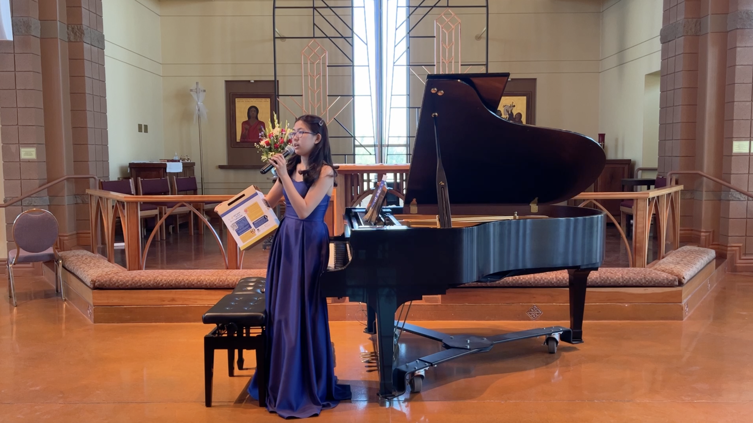 A woman in a long blue dress singing into a microphone while playing a grand piano in a church or concert hall.