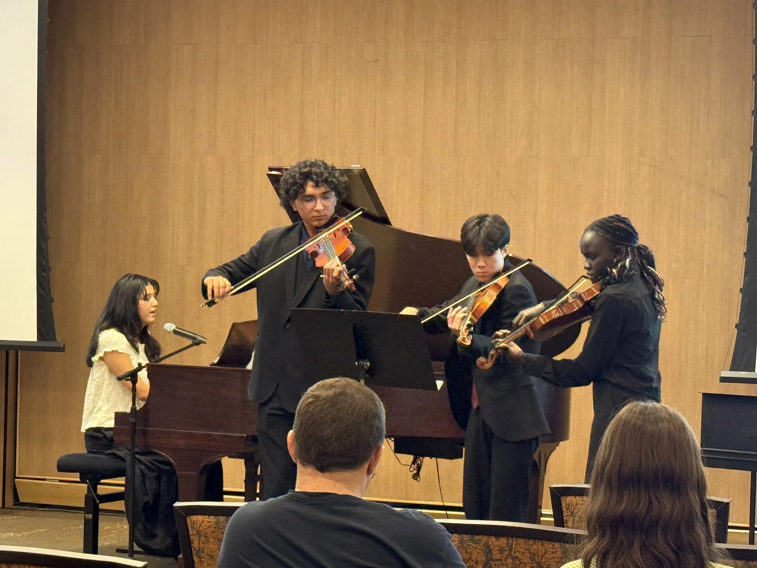 Four musicians performing on stage: a woman playing the piano, and three others playing violins, with an audience watching in the foreground.