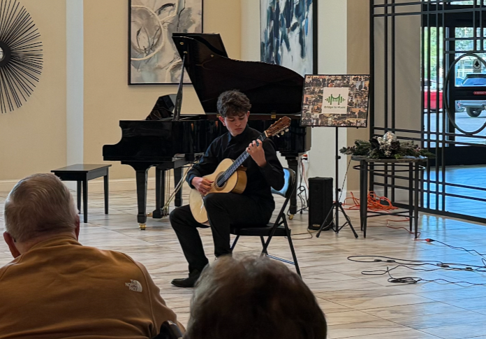 Young man playing classical guitar in a performance room with a grand piano, art on the walls, and a small audience watching.