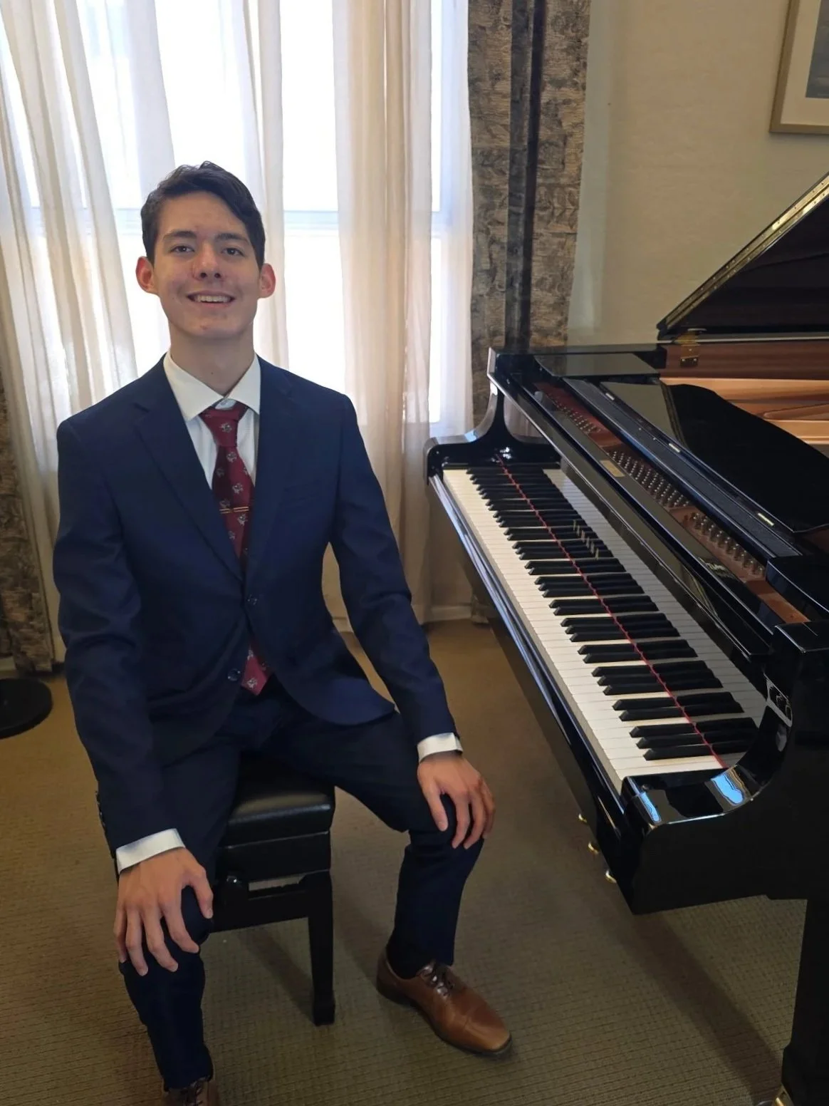 Young man in a blue suit sitting on a piano bench next to a grand piano in a room with cream-colored curtains.