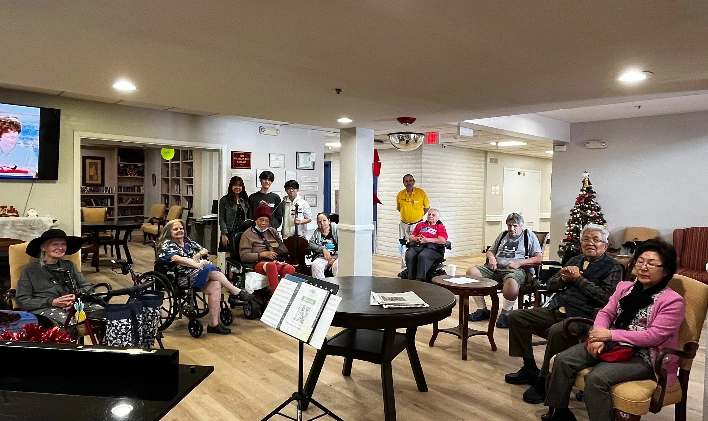 Group of seniors and young people gathered in a community room, some in wheelchairs, with a Christmas tree, music stand, and decorations, suggesting a holiday celebration or social event.