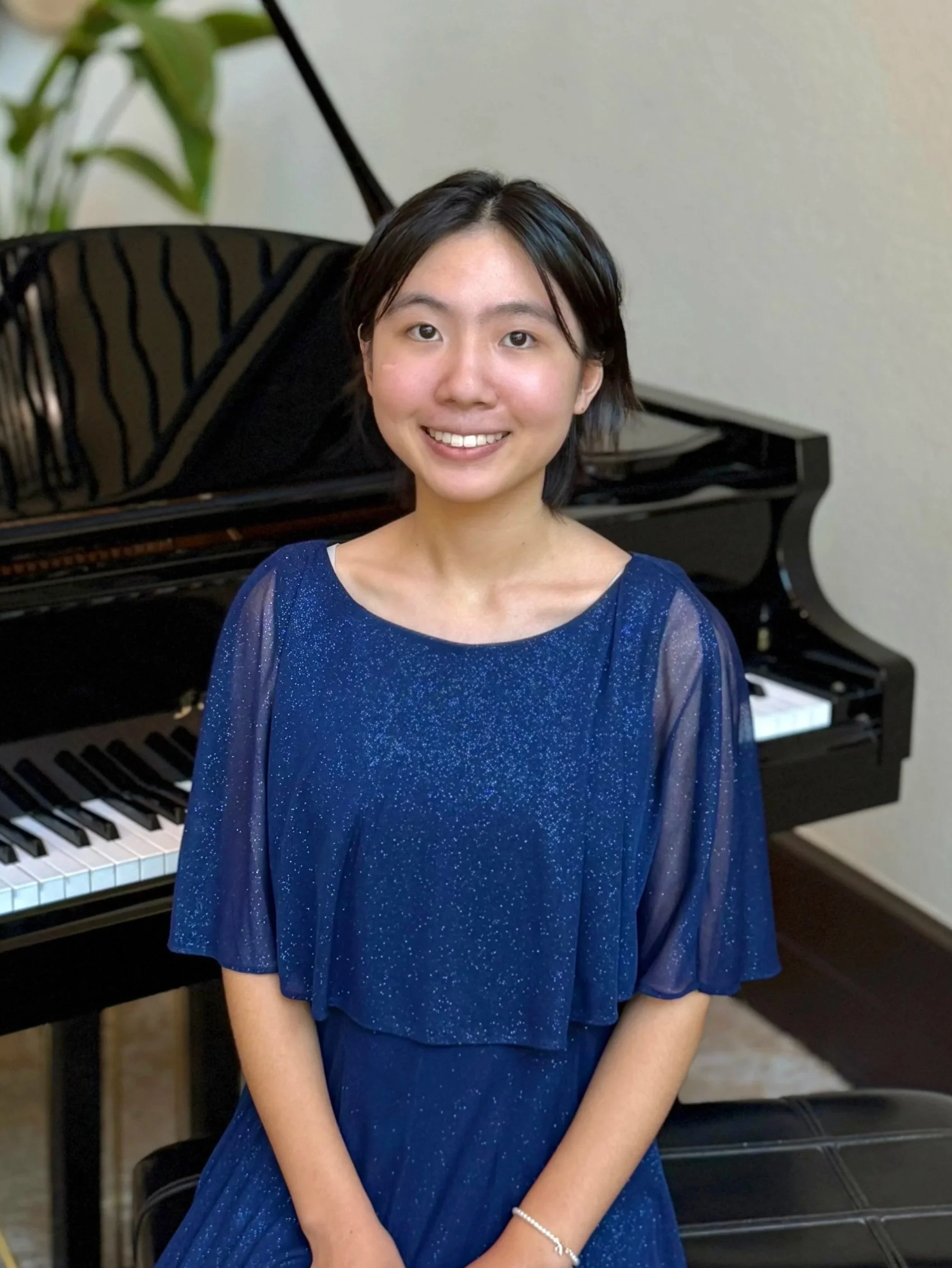 A young woman in a blue, sparkly dress sitting in front of a grand piano, smiling at the camera.